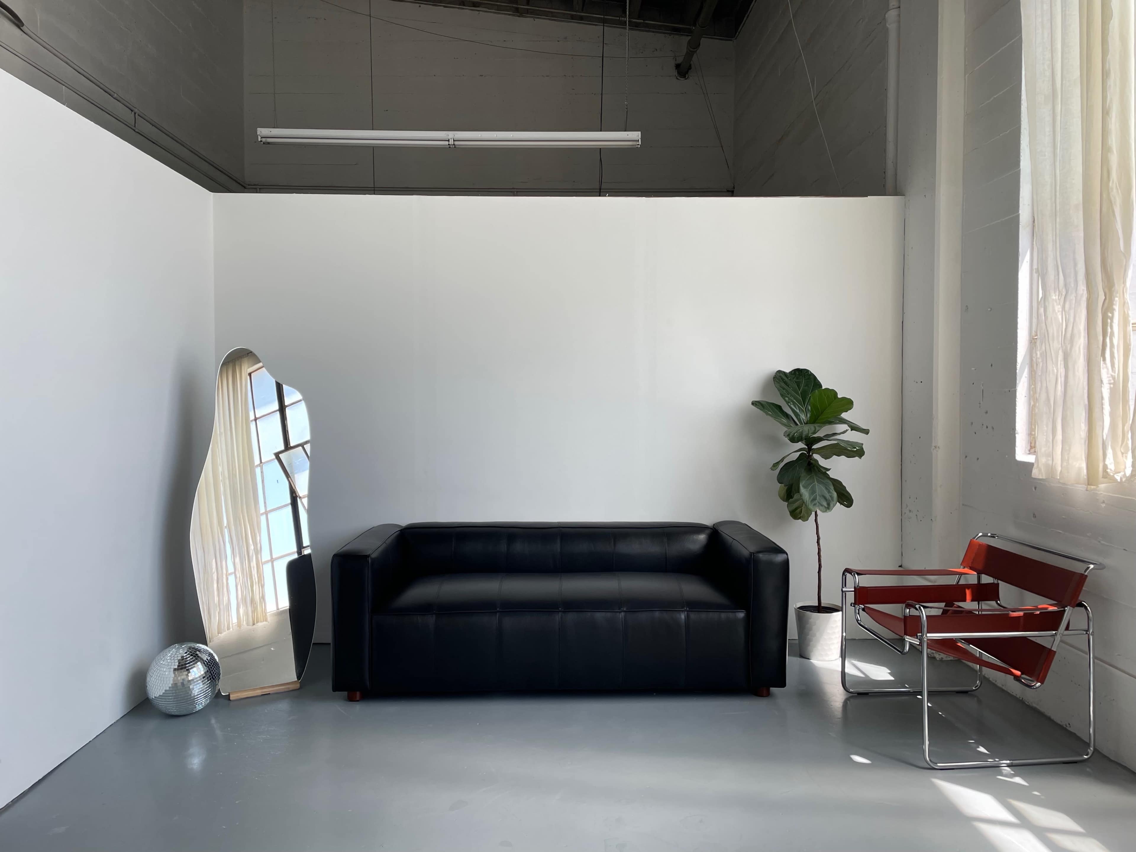The image shows a black leather sofa positioned against a white wall, accompanied by a large mirror, a potted plant, and a red-framed chair.