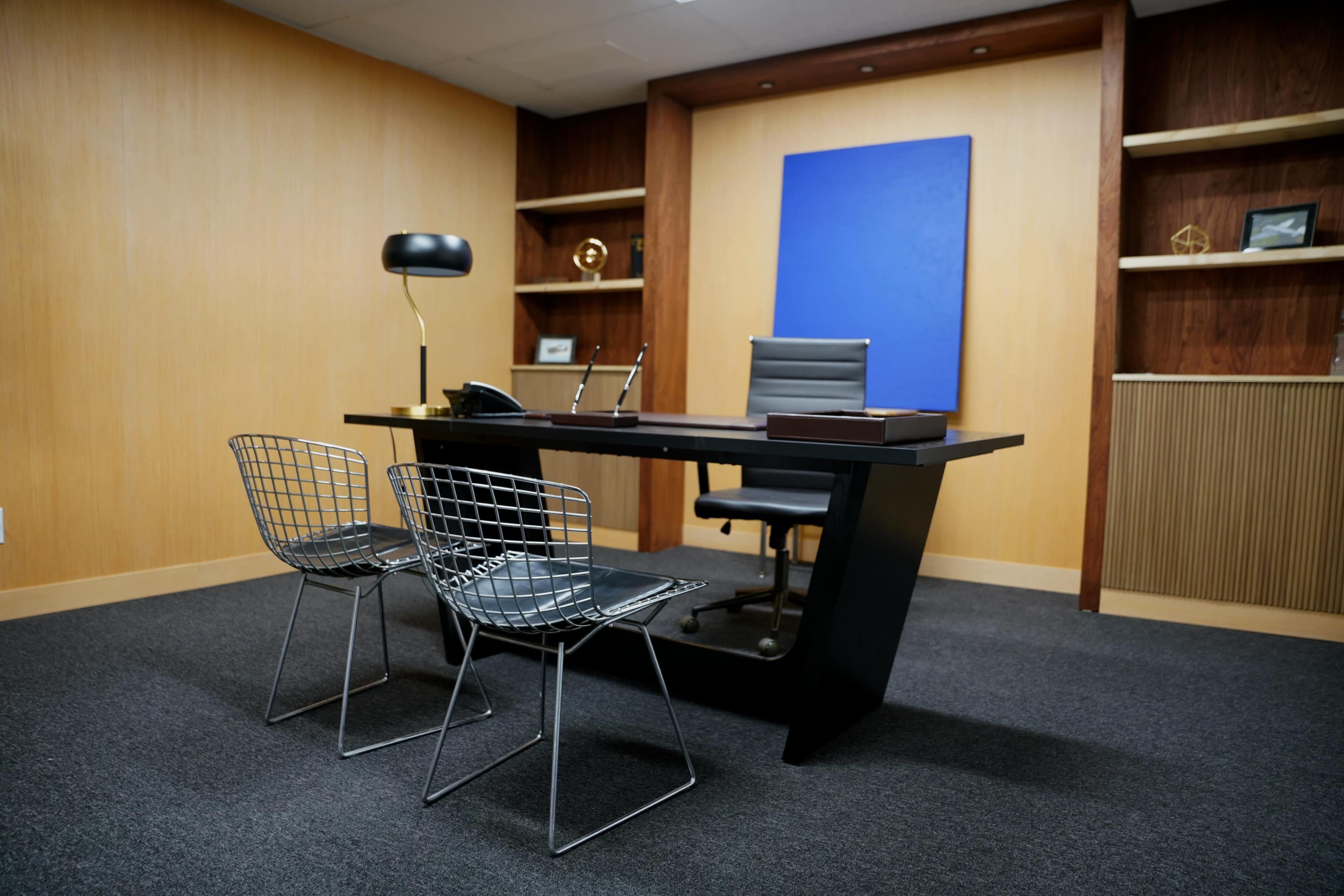 The image shows a minimalist office space featuring a black desk, a chair, and two wire chairs, with wooden shelves and a blue art piece on the wall.