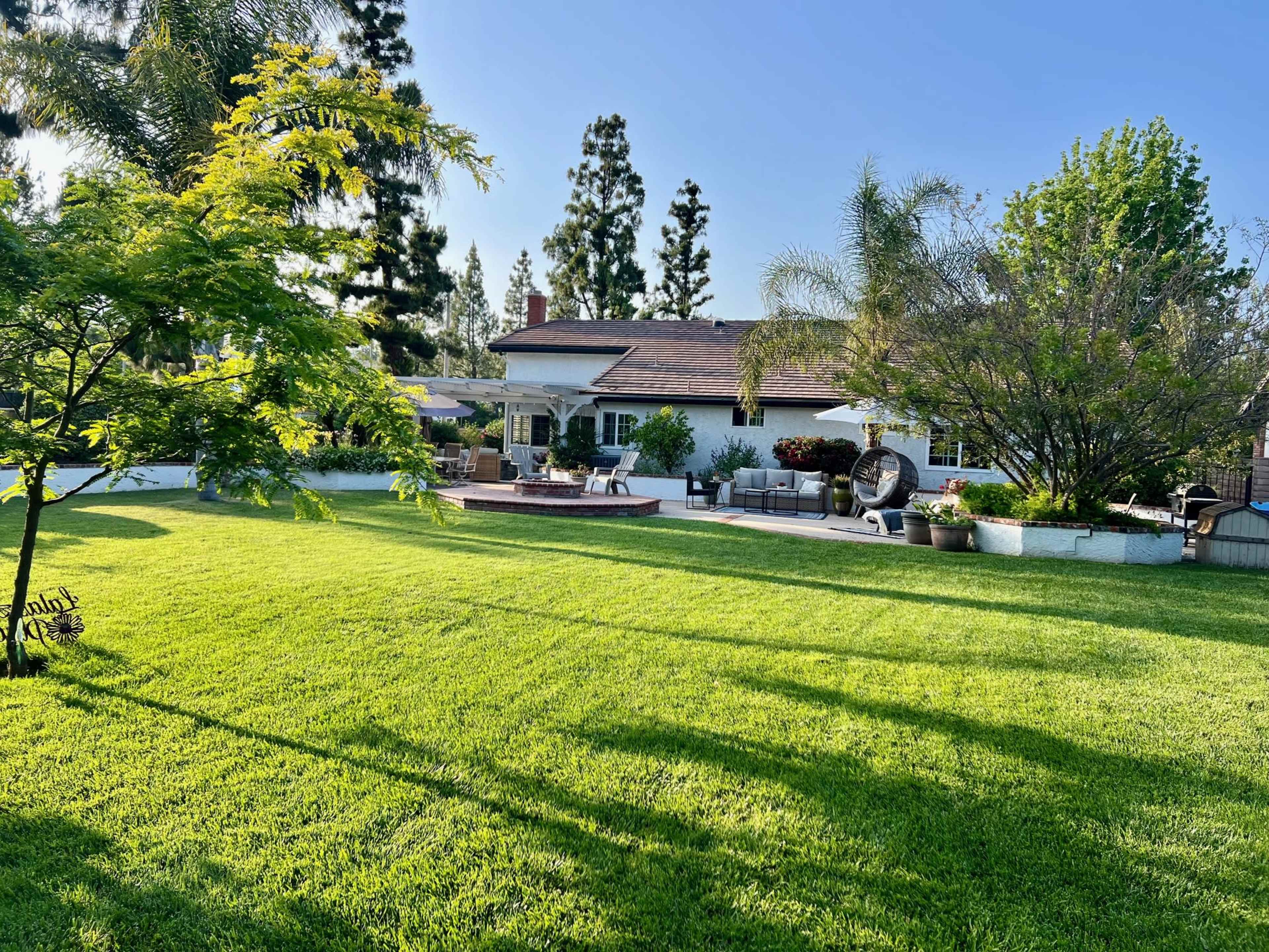 A well-maintained lawn and landscaped backyard surround a single-story home with outdoor seating and trees.