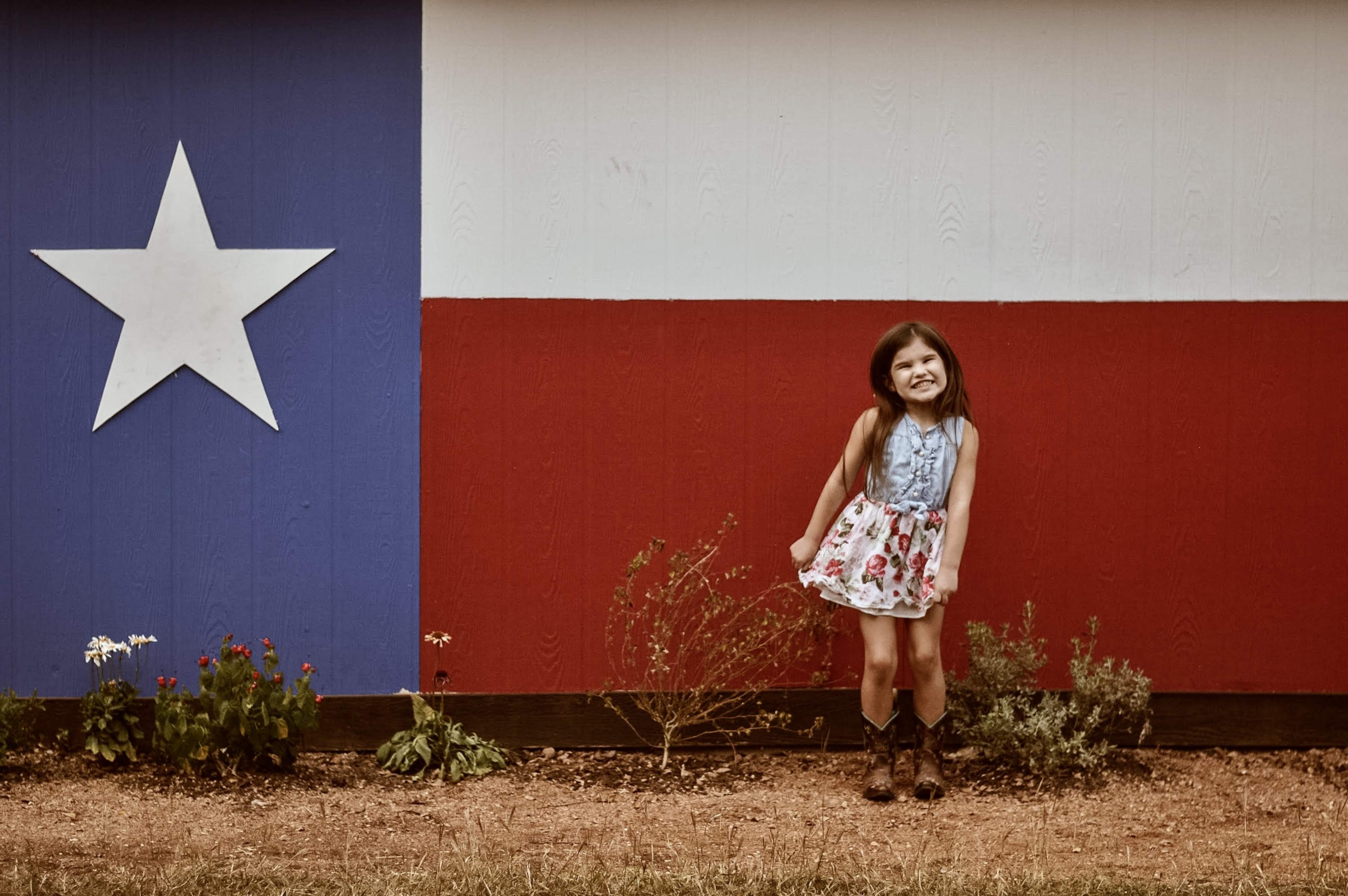 A girl in a floral dress stands in front of a wall painted with the Texas flag.