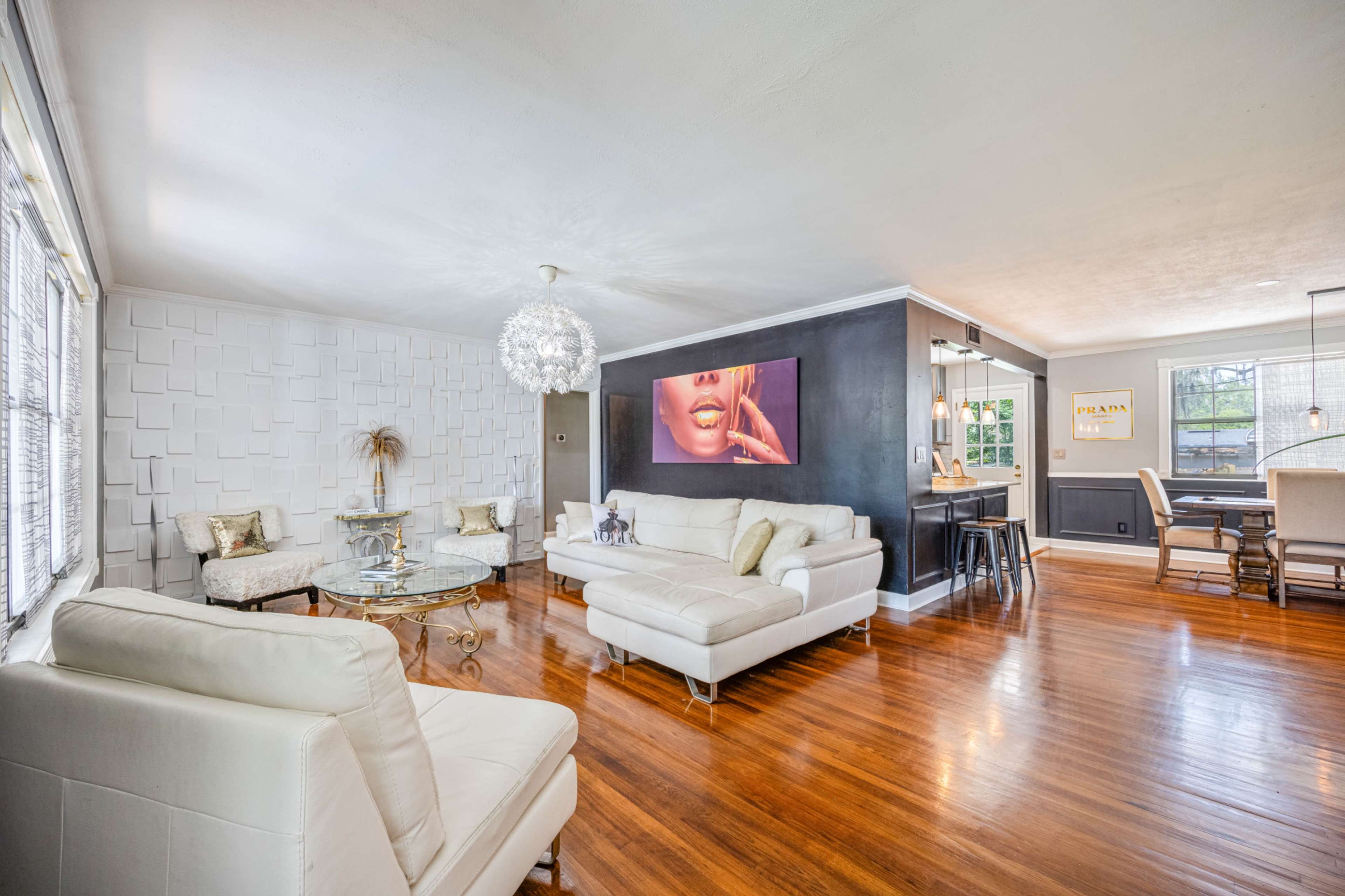 The living room features a light-colored sectional sofa, modern chairs, a glass coffee table, and a large wall-mounted artwork, with hardwood floors and natural light coming through the windows.