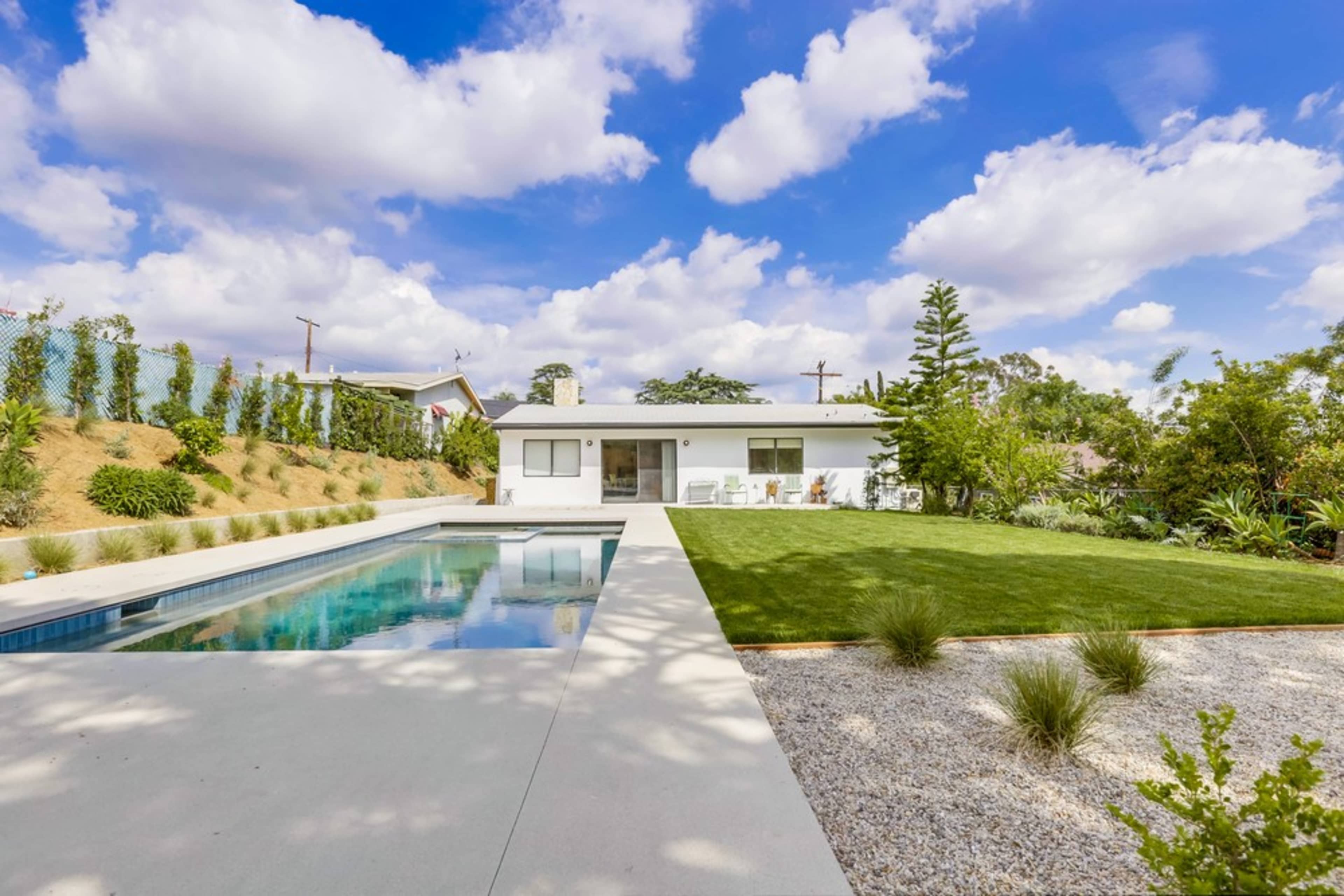 A modern house with a pool in the foreground, set in a landscaped yard that features green grass and decorative plants under a partly cloudy sky.