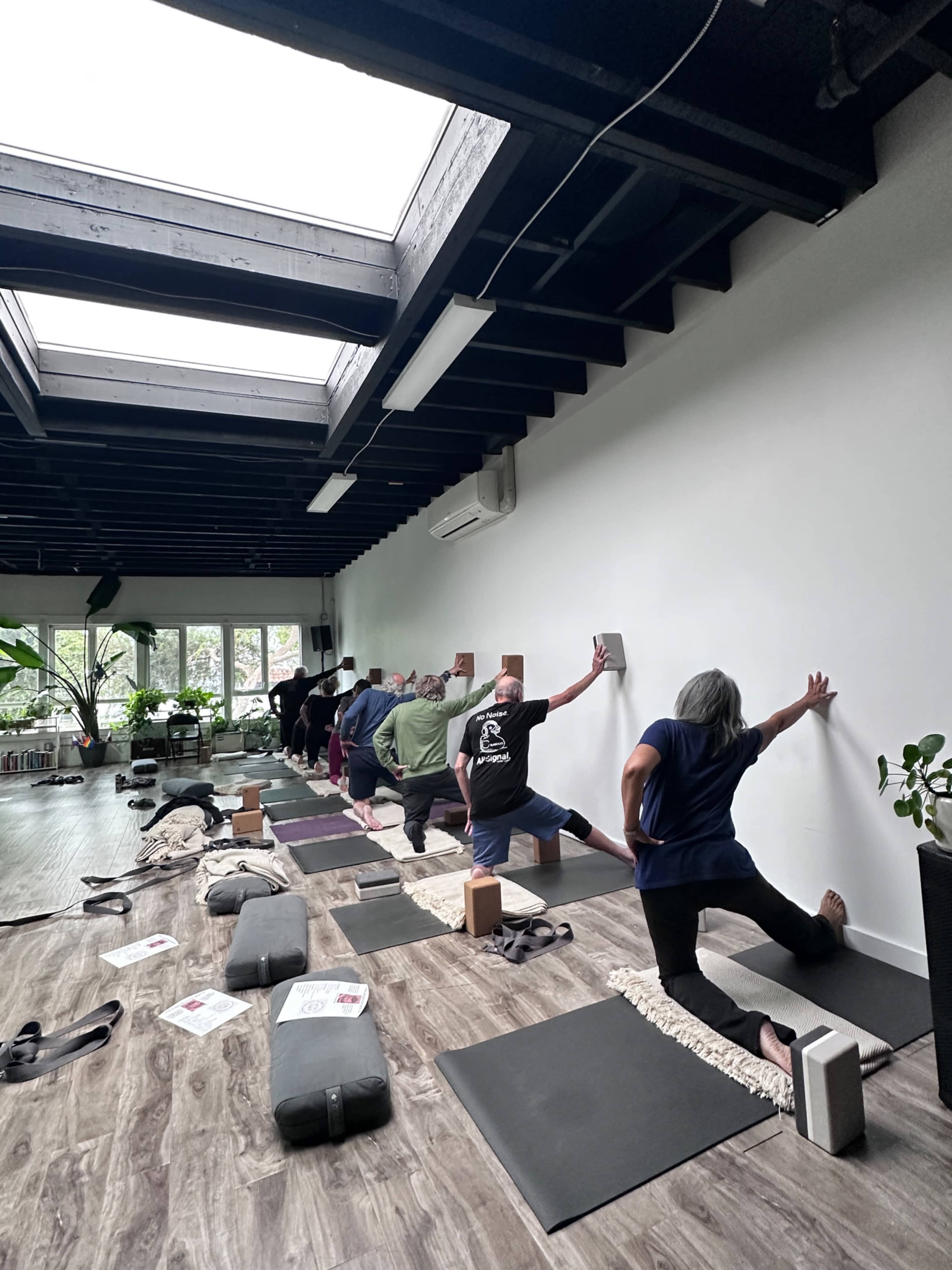A group of people practice yoga in a studio with a bright skylight and various props scattered on the floor.