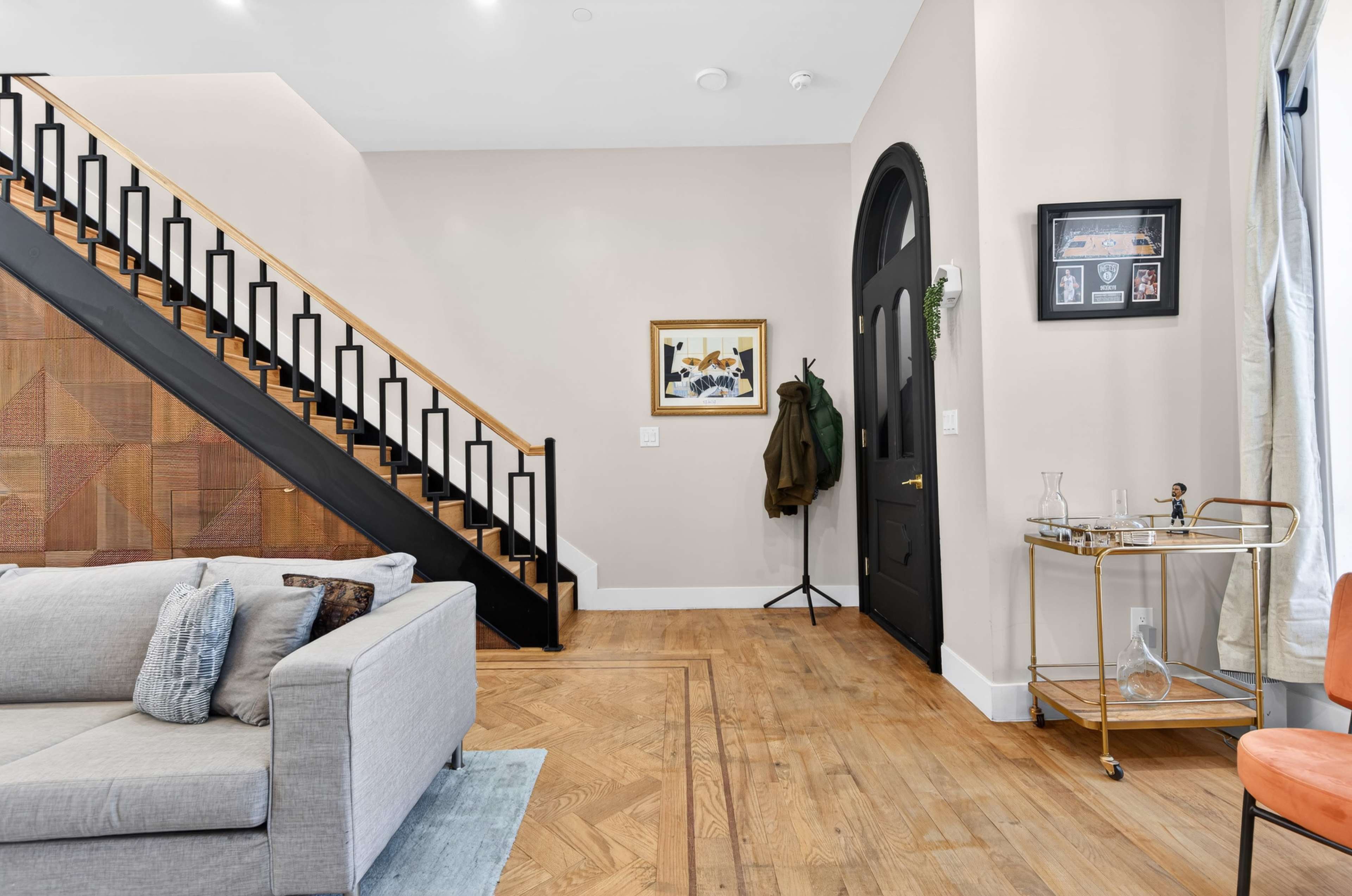 The image shows a modern living space with a staircase leading to an upper level, framed by a coat rack and a small bar cart in the corner.