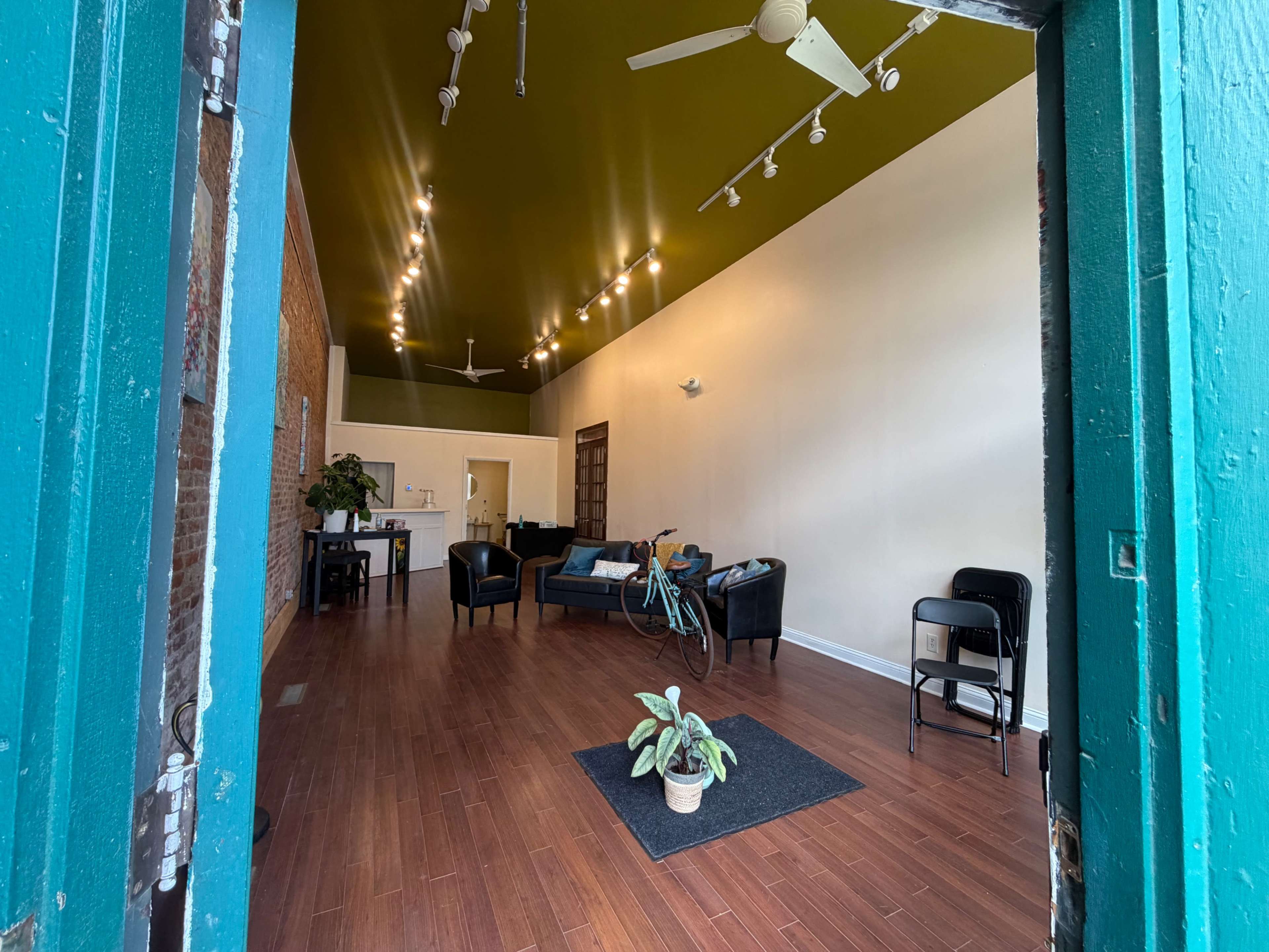 The image shows the interior of an empty, well-lit space with a green ceiling, wooden flooring, and minimal furniture, including chairs, sofas, and a small plant.