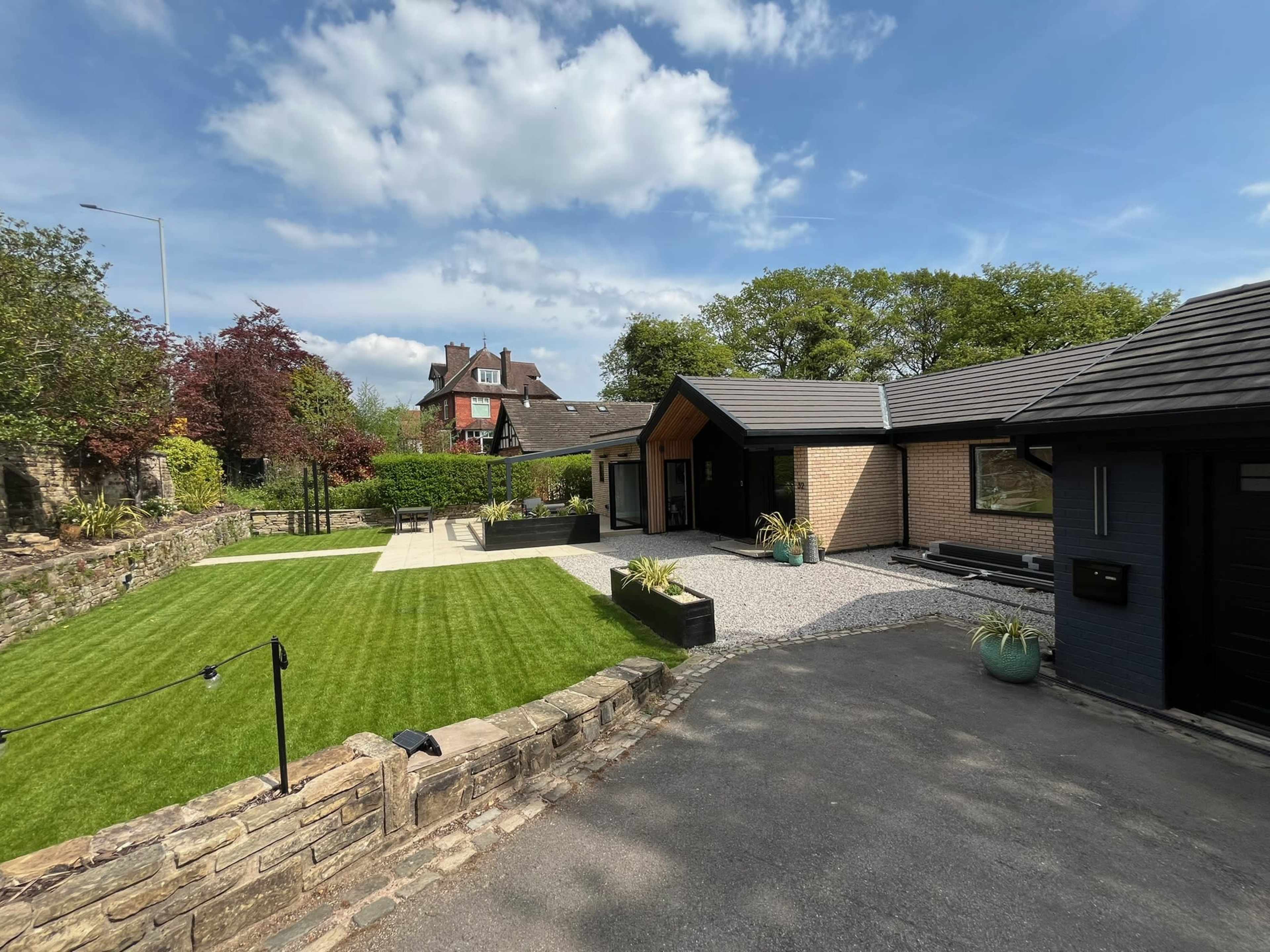 The image shows a modern, single-story house with a landscaped lawn, paved walkways, and a combination of stone and wood exterior finishes under a blue sky with some clouds.