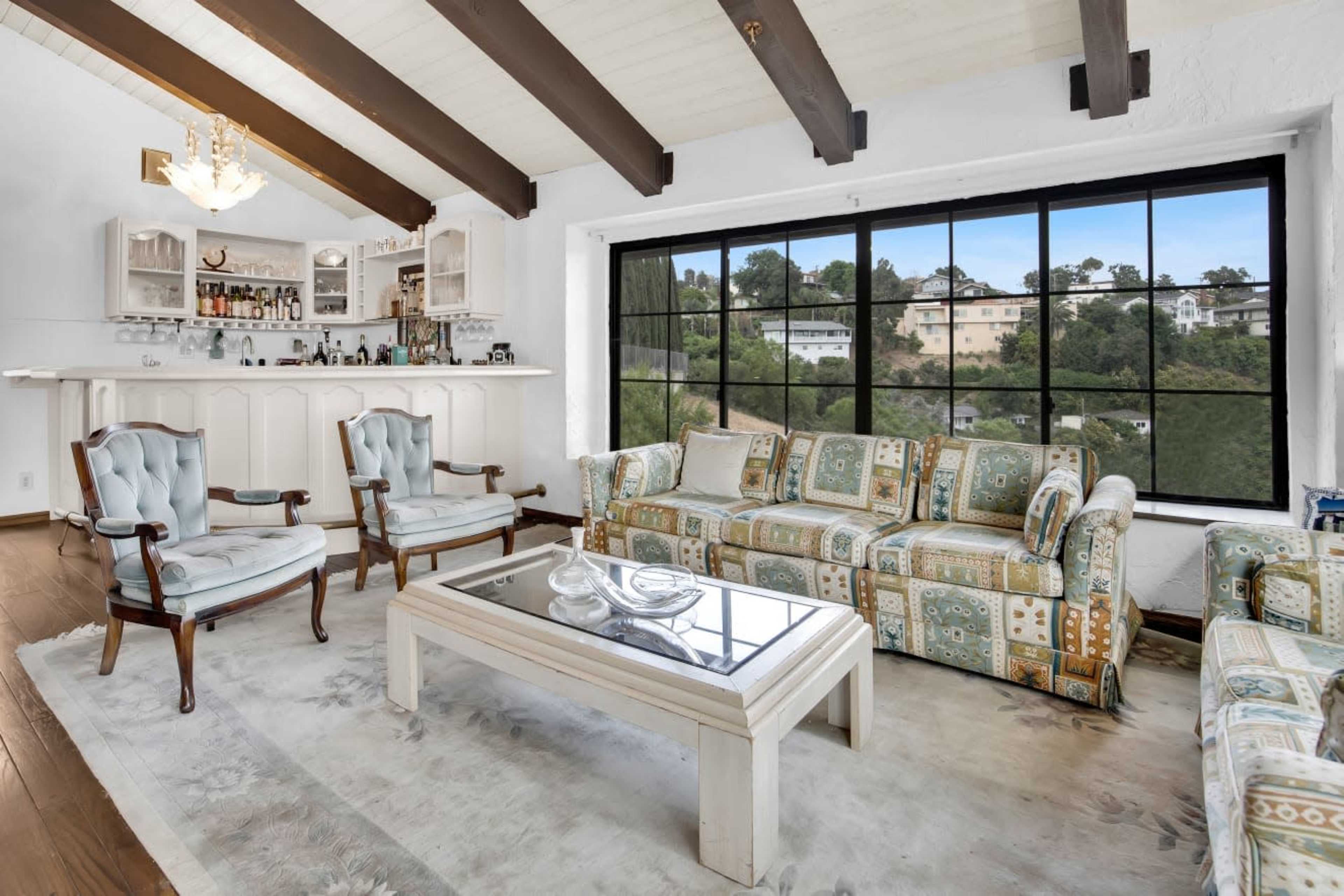 The living room features a bar area, a patterned sofa set, and large windows overlooking a green hillside.
