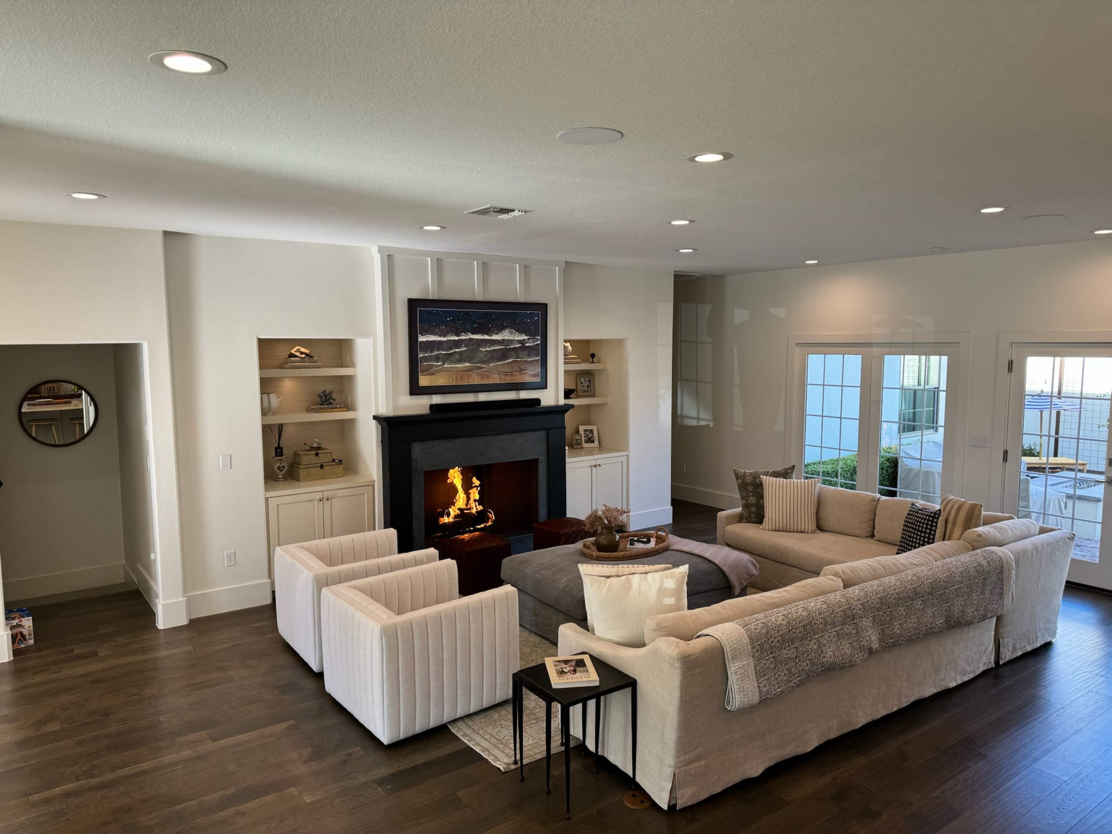 A modern living room with a large sectional sofa, two armchairs, a fireplace, built-in shelves, and large windows offering views of the outdoors.