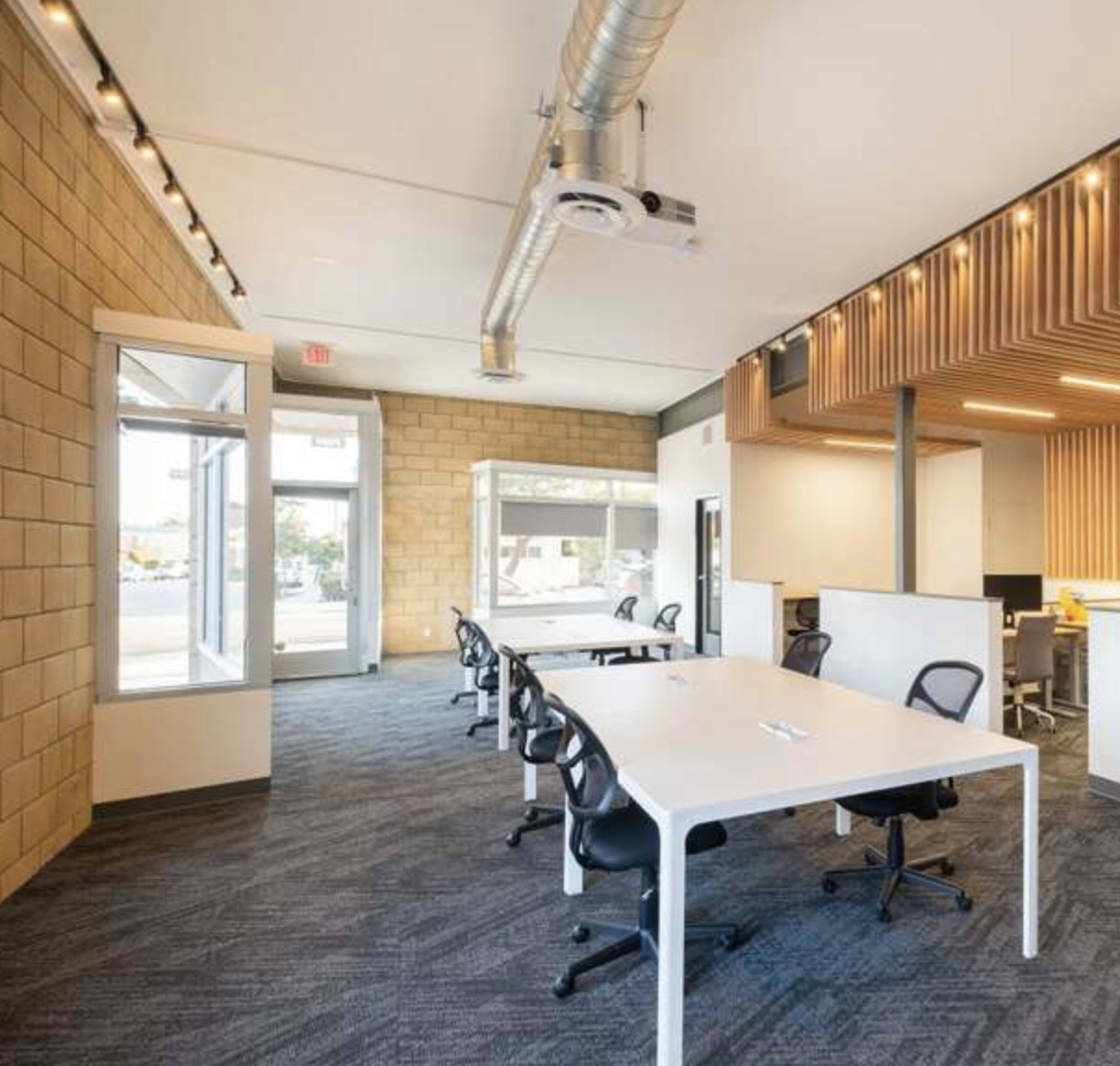A modern office space featuring several white desks with black chairs, large windows, and a mix of wood and concrete wall textures.