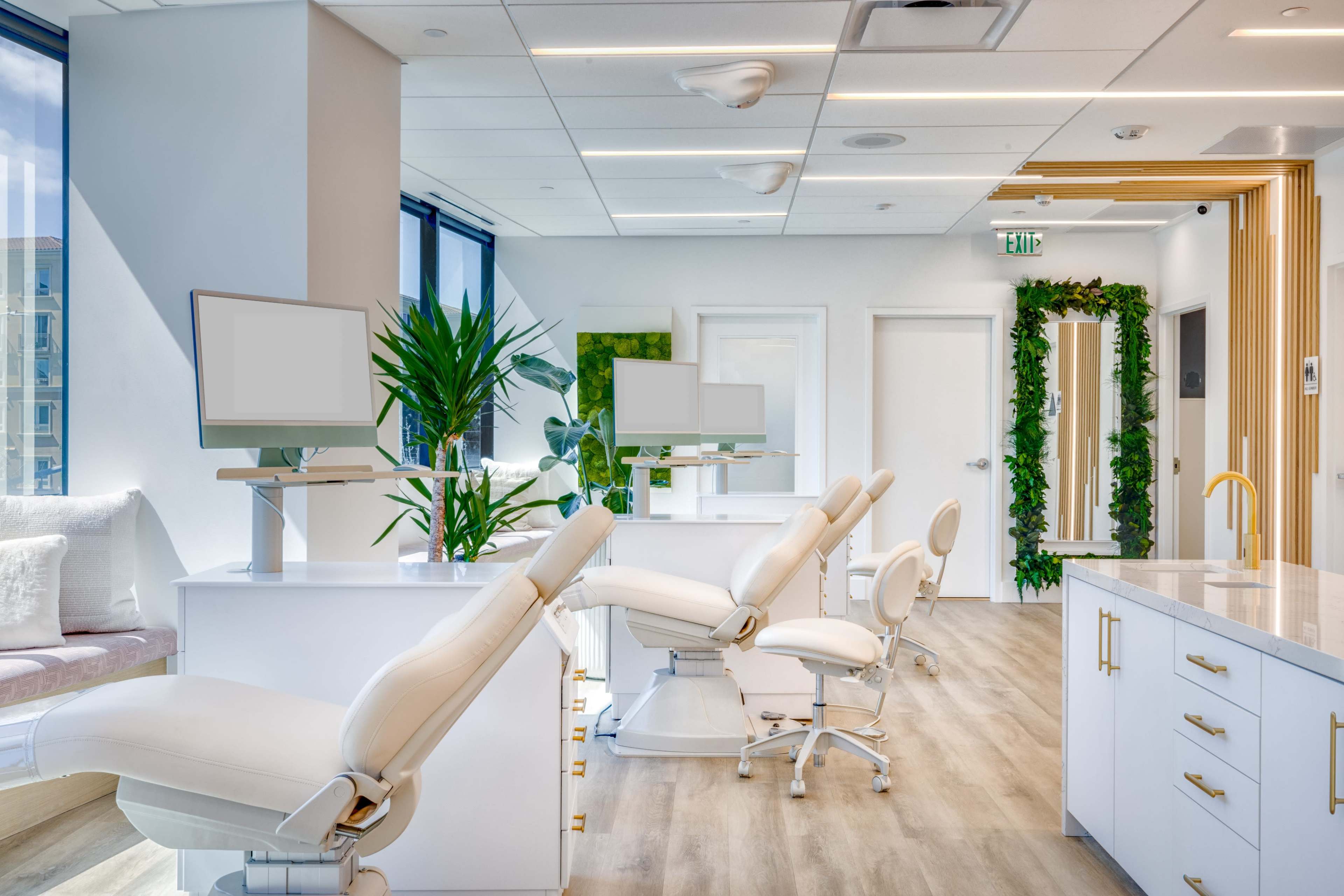 The image shows a modern dental office with several white dental chairs, computers on white desks, and green plants in a well-lit space.