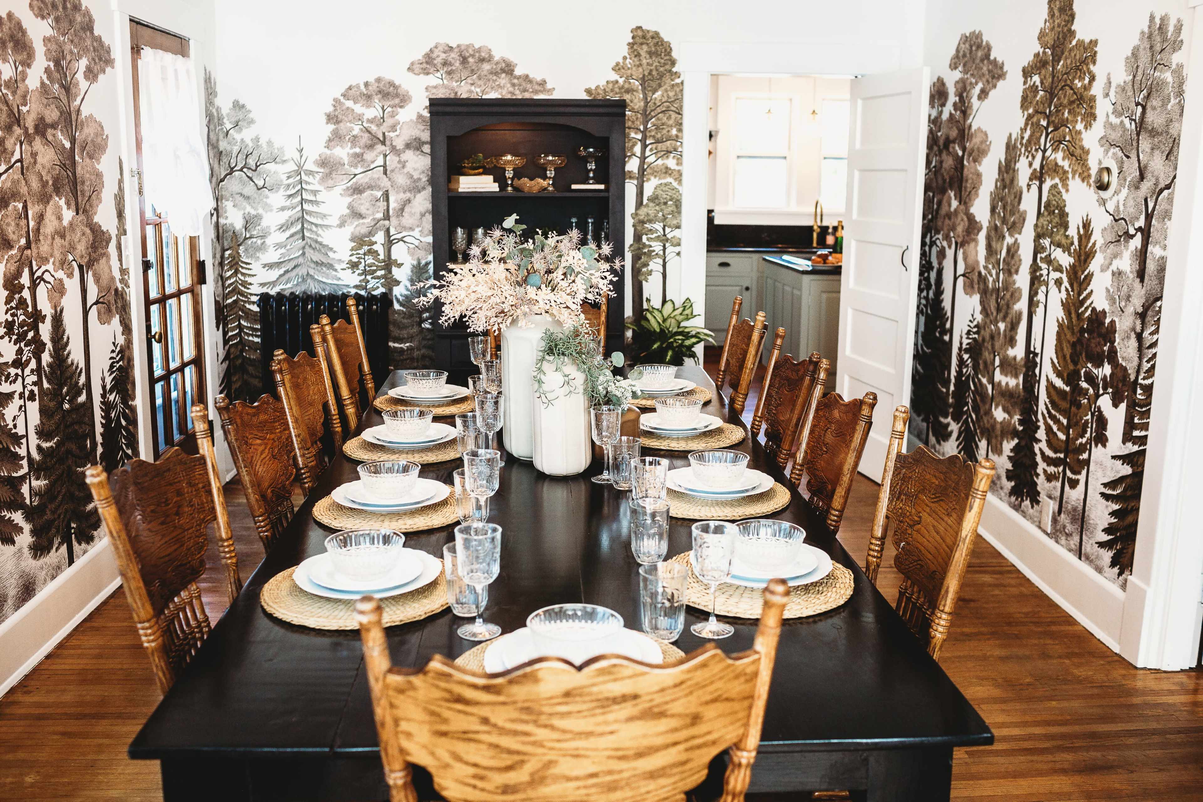 A large dining table is set for a meal, surrounded by wooden chairs and featuring wallpaper with a nature-inspired landscape.