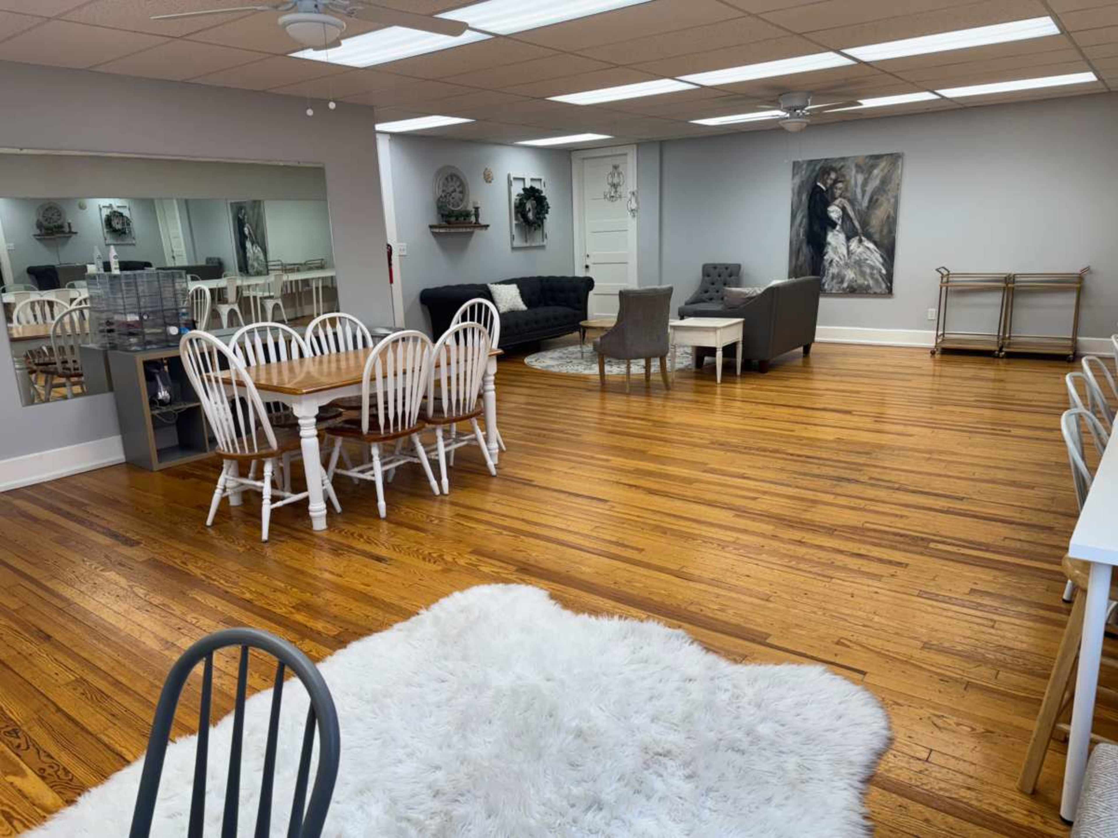 The image shows a spacious, well-lit room with wooden floors, featuring a dining table with white chairs, a grey sofa, various seating arrangements, and a decorative wall painting.