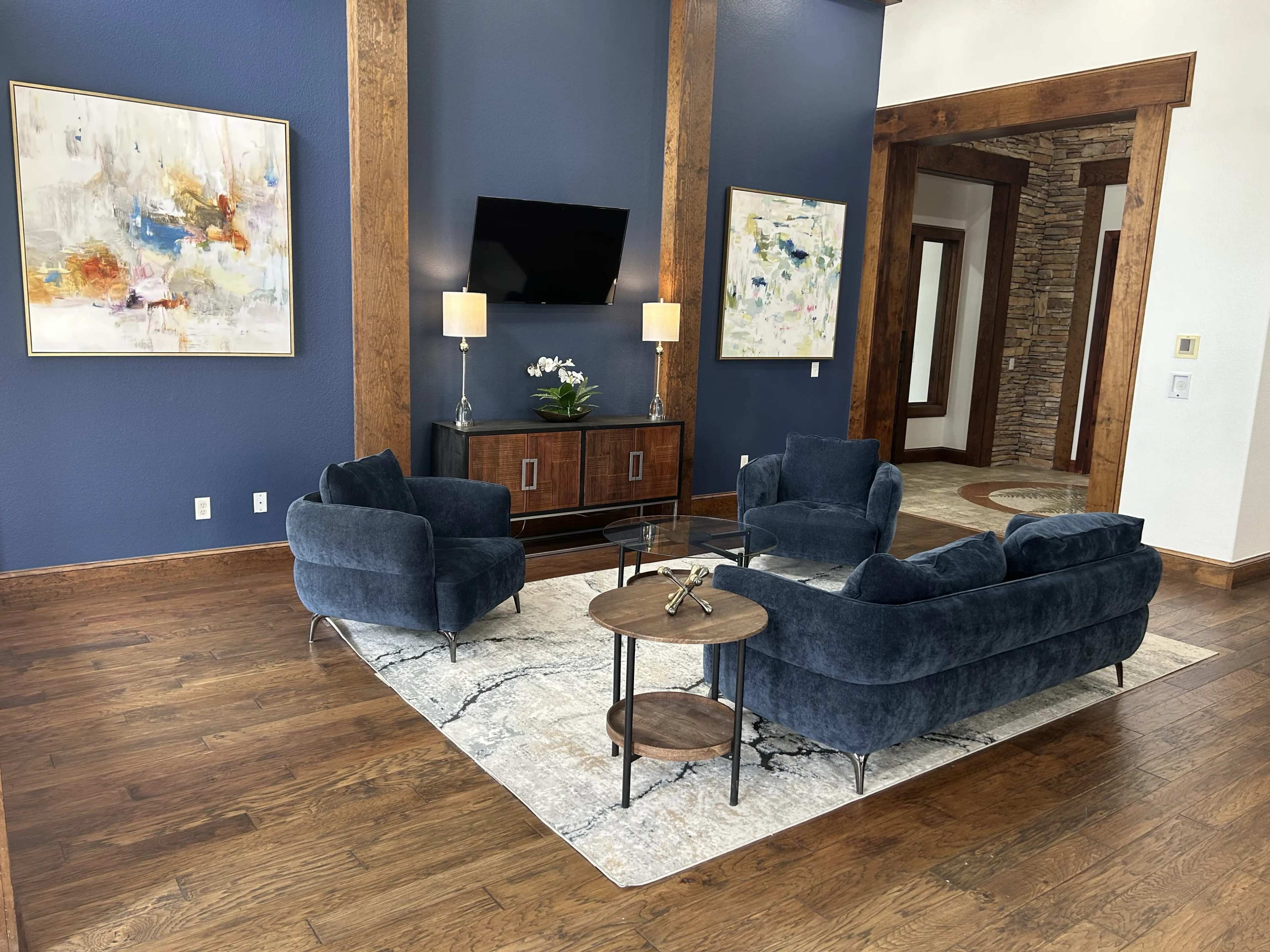 A stylish living room features two blue velvet armchairs and a matching sofa arranged around a circular coffee table on a patterned area rug, with a large abstract painting and a television mounted on the wall.