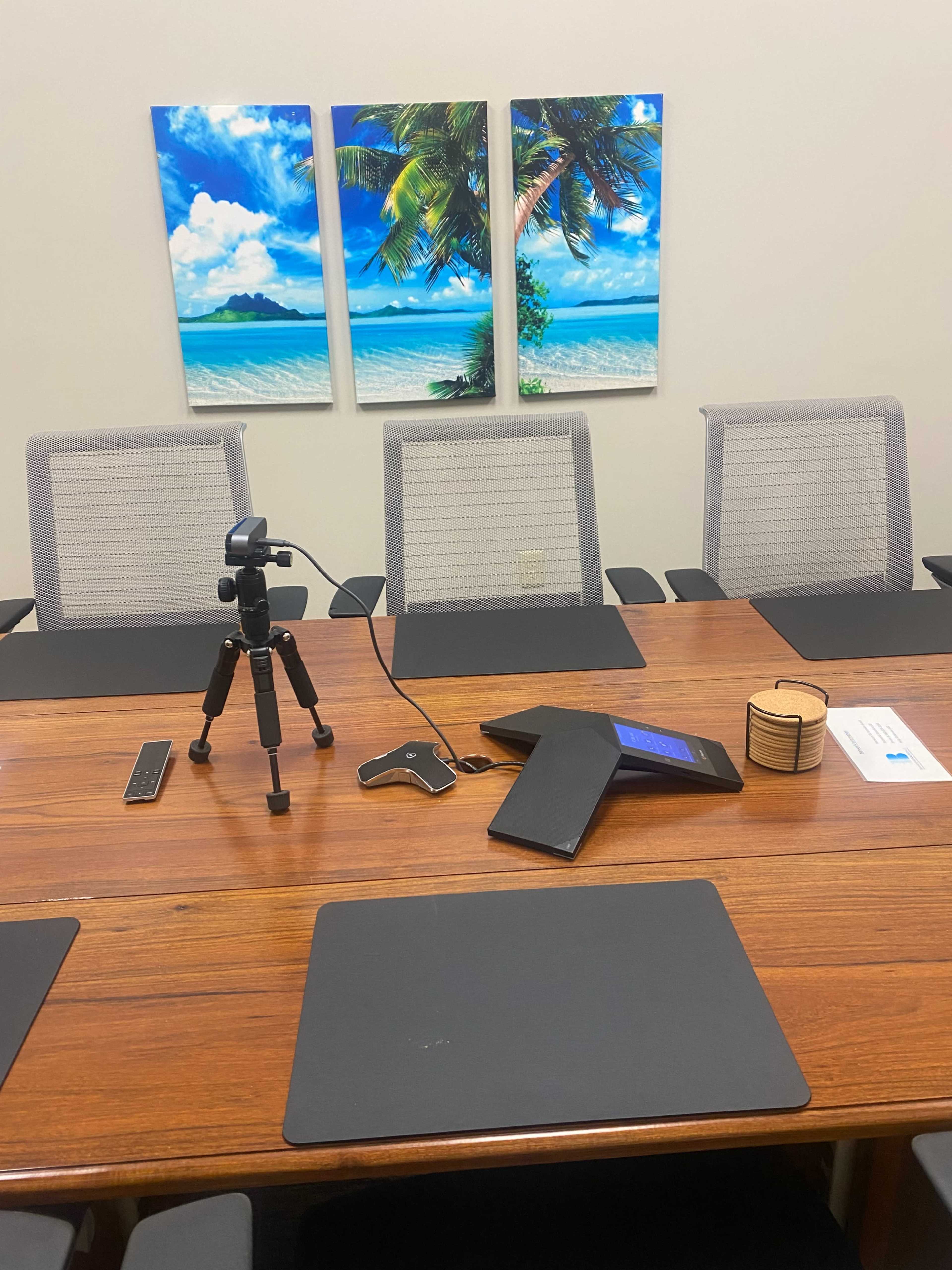 A conference table is set with a tripod, remote control, and a tablet, surrounded by several empty chairs, with beach-themed artwork on the wall behind.