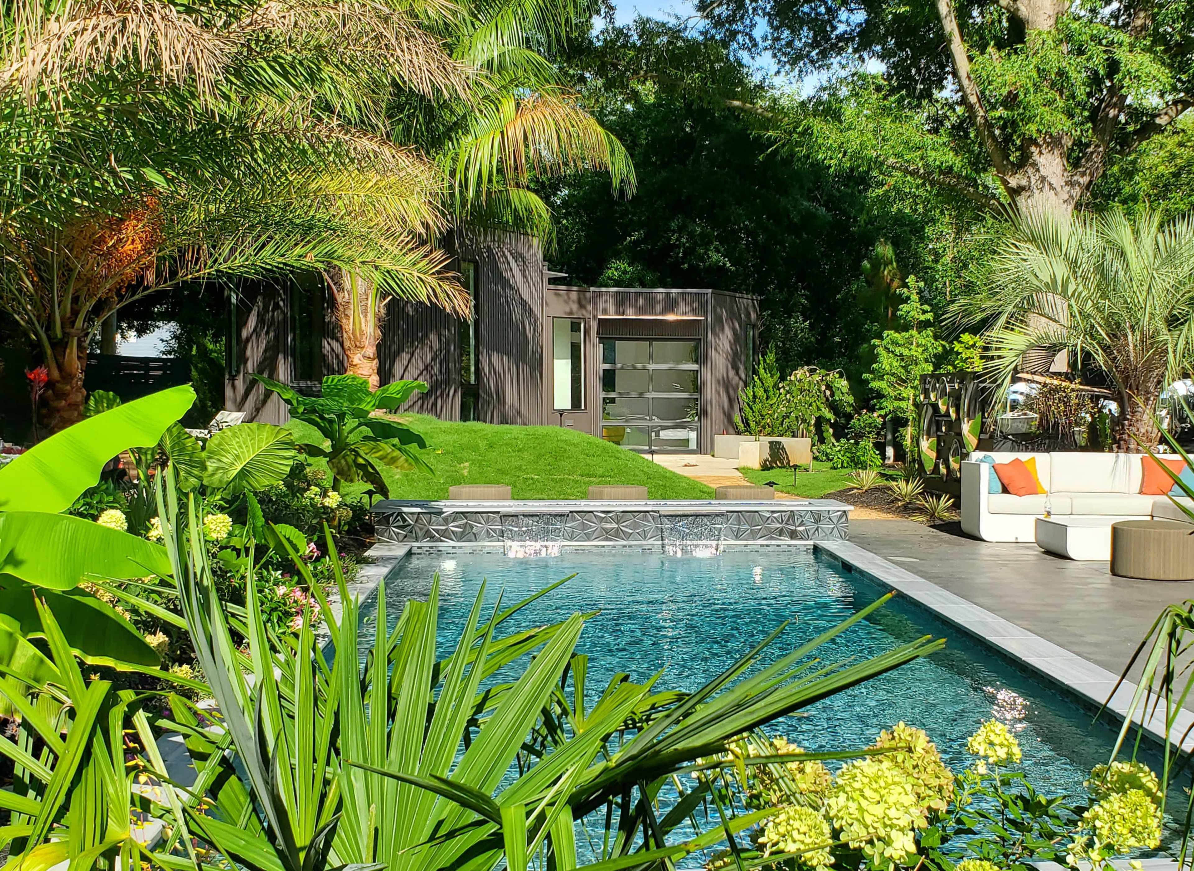 The image shows a landscaped backyard featuring a swimming pool surrounded by lush greenery and modern outdoor furniture.