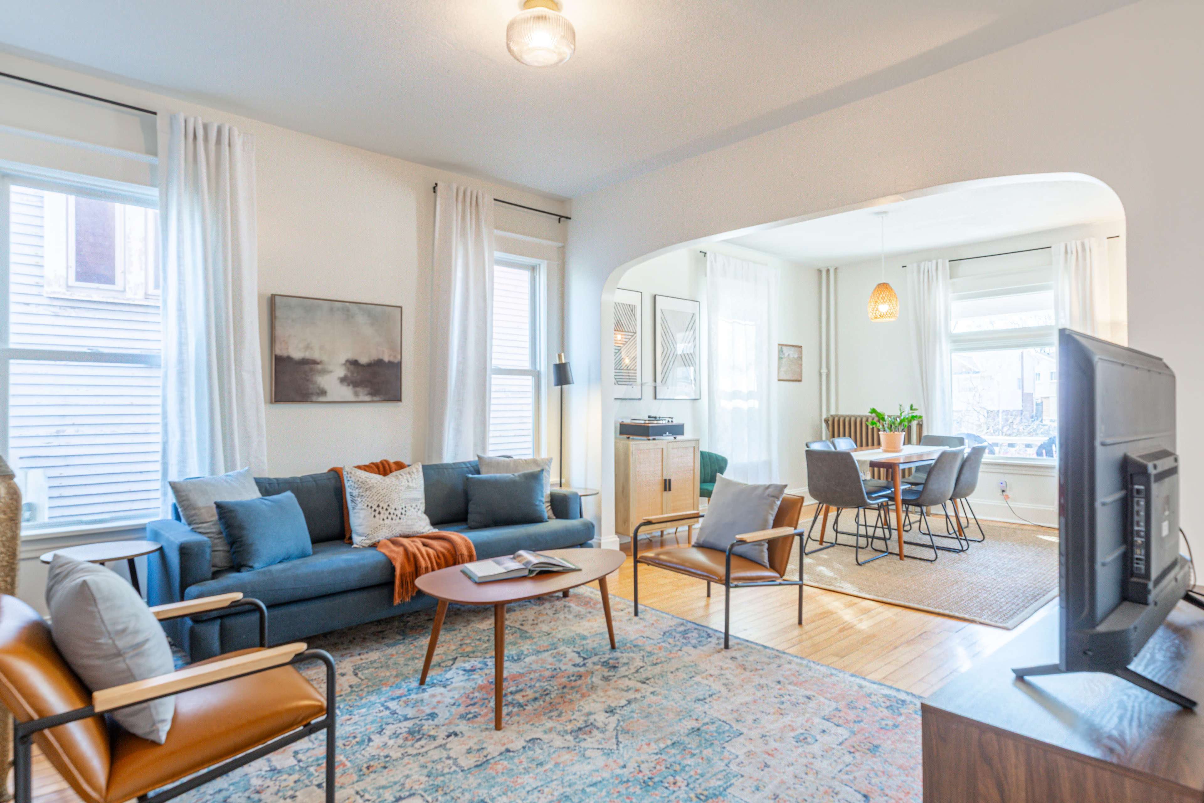 The image shows a cozy living room with a blue sectional sofa, a wooden coffee table, and a dining area featuring a round table and chairs, all illuminated by natural light from large windows.