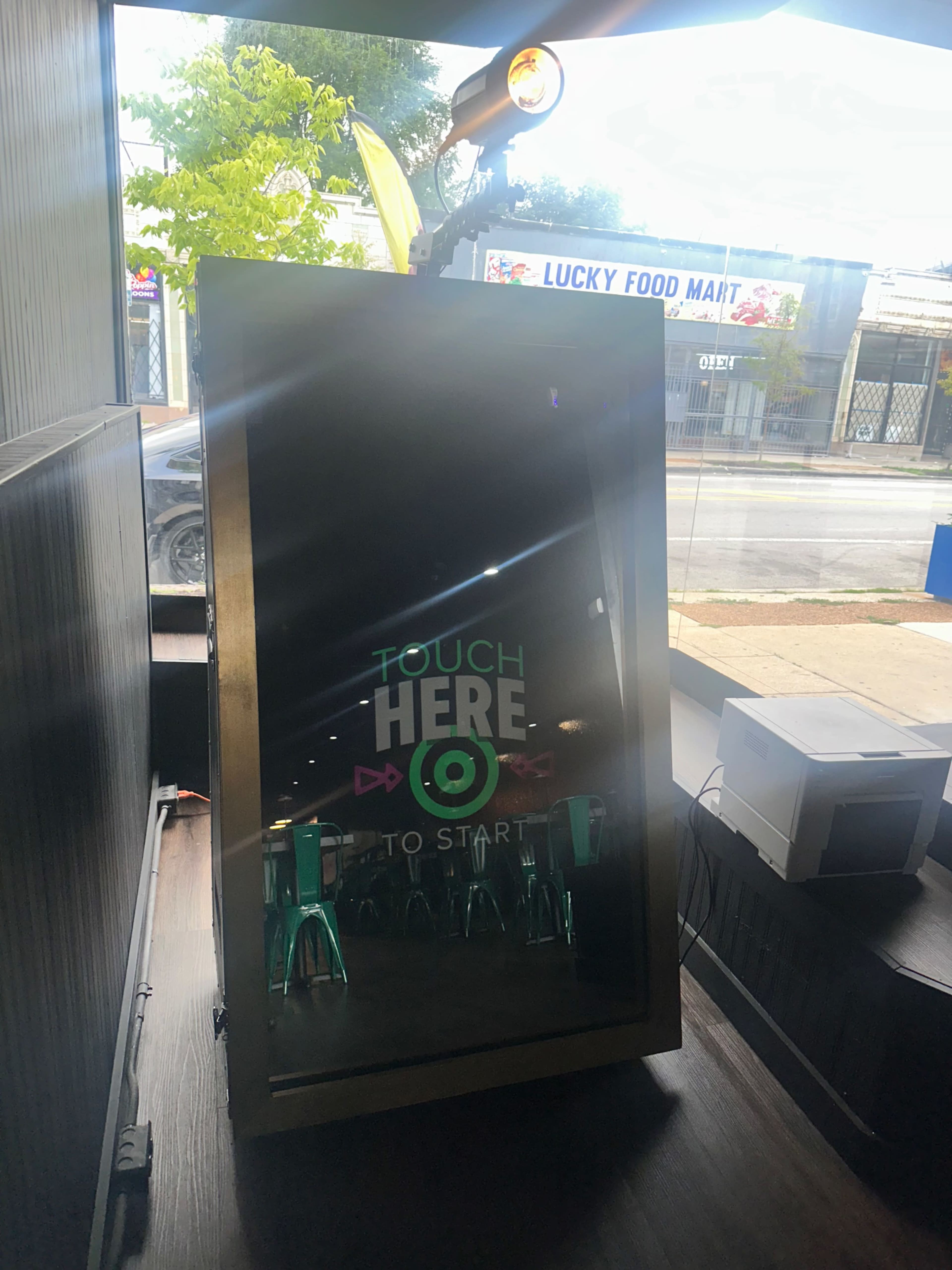 A touchscreen kiosk with the words "TOUCH HERE TO START" is positioned in a well-lit area near a window, with chairs visible in front.