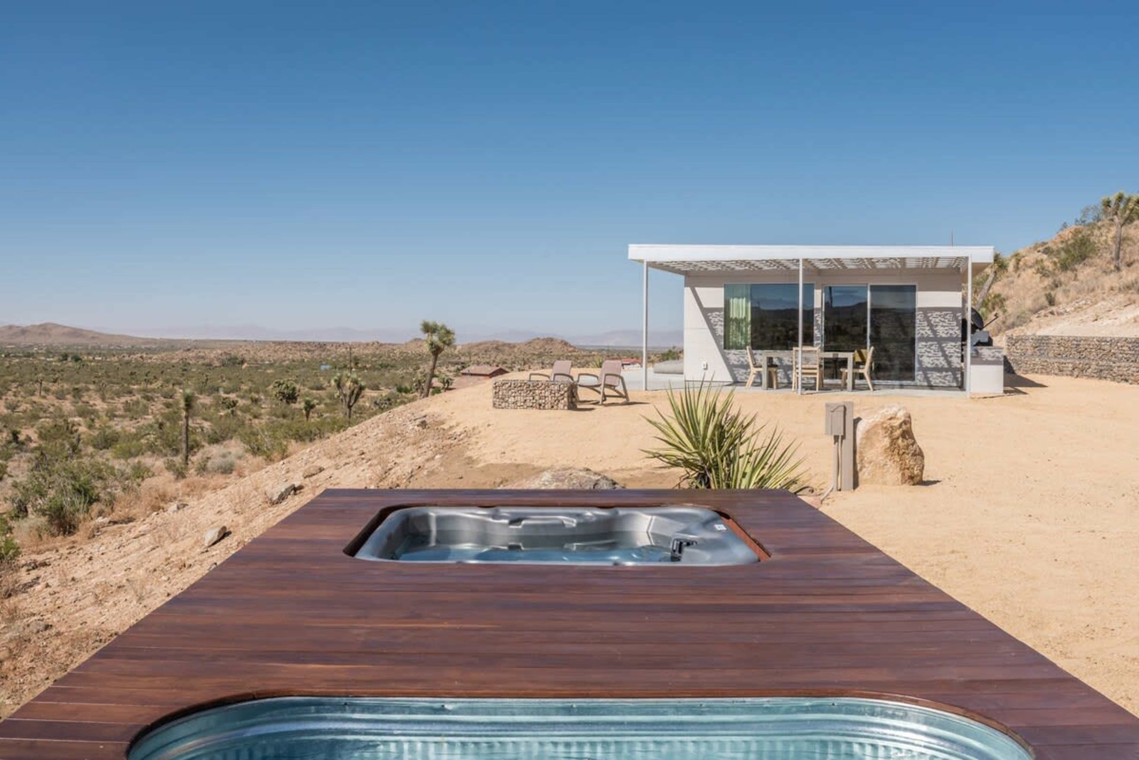 A modern house with large windows sits isolated on a rocky hillside, alongside a hot tub overlooking a desert landscape.