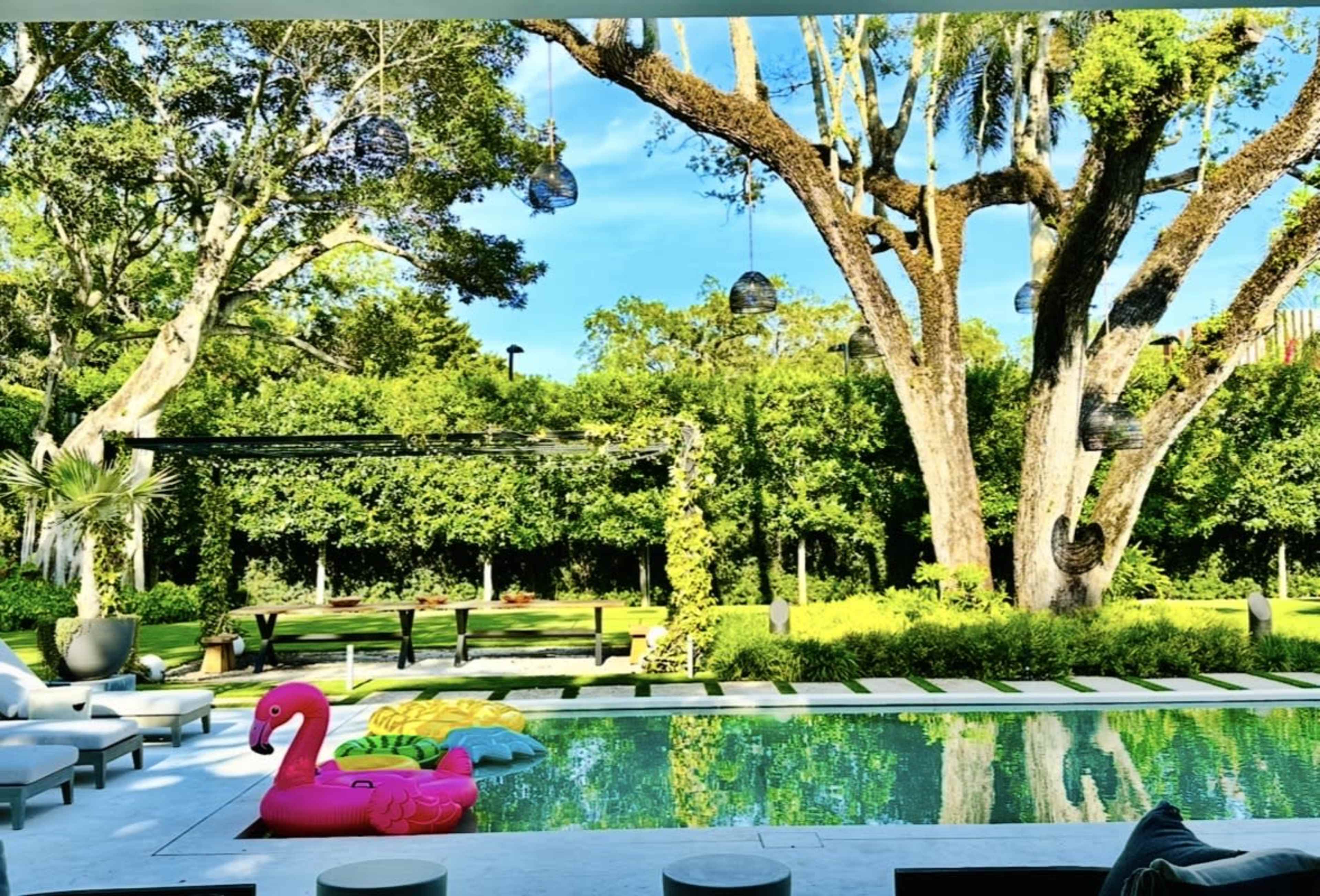 A bright, landscaped garden features a swimming pool with a pink flamingo float, surrounded by lush greenery and large trees.
