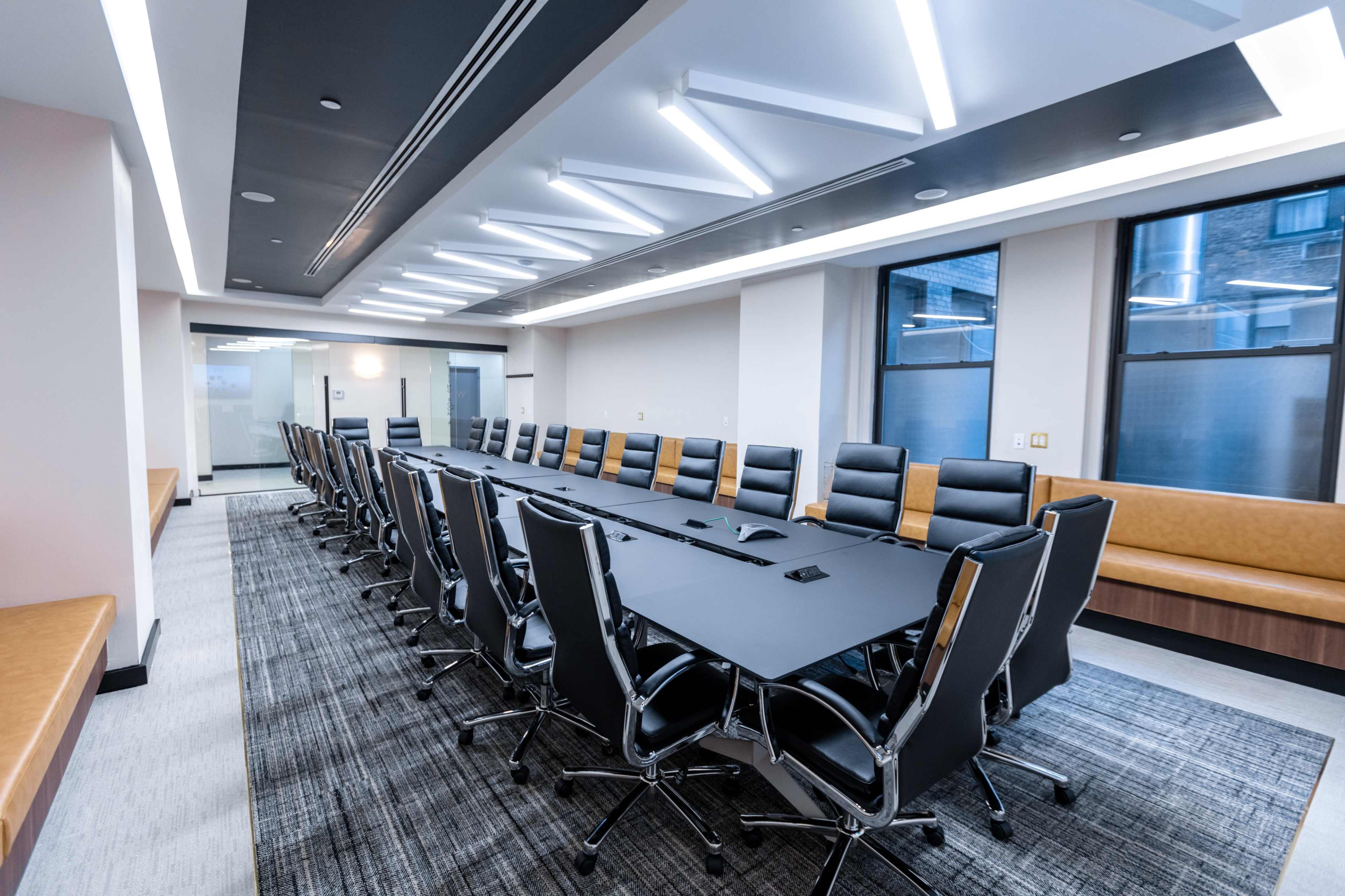 A modern conference room featuring a long black table surrounded by black leather chairs, with large windows and overhead lighting.
