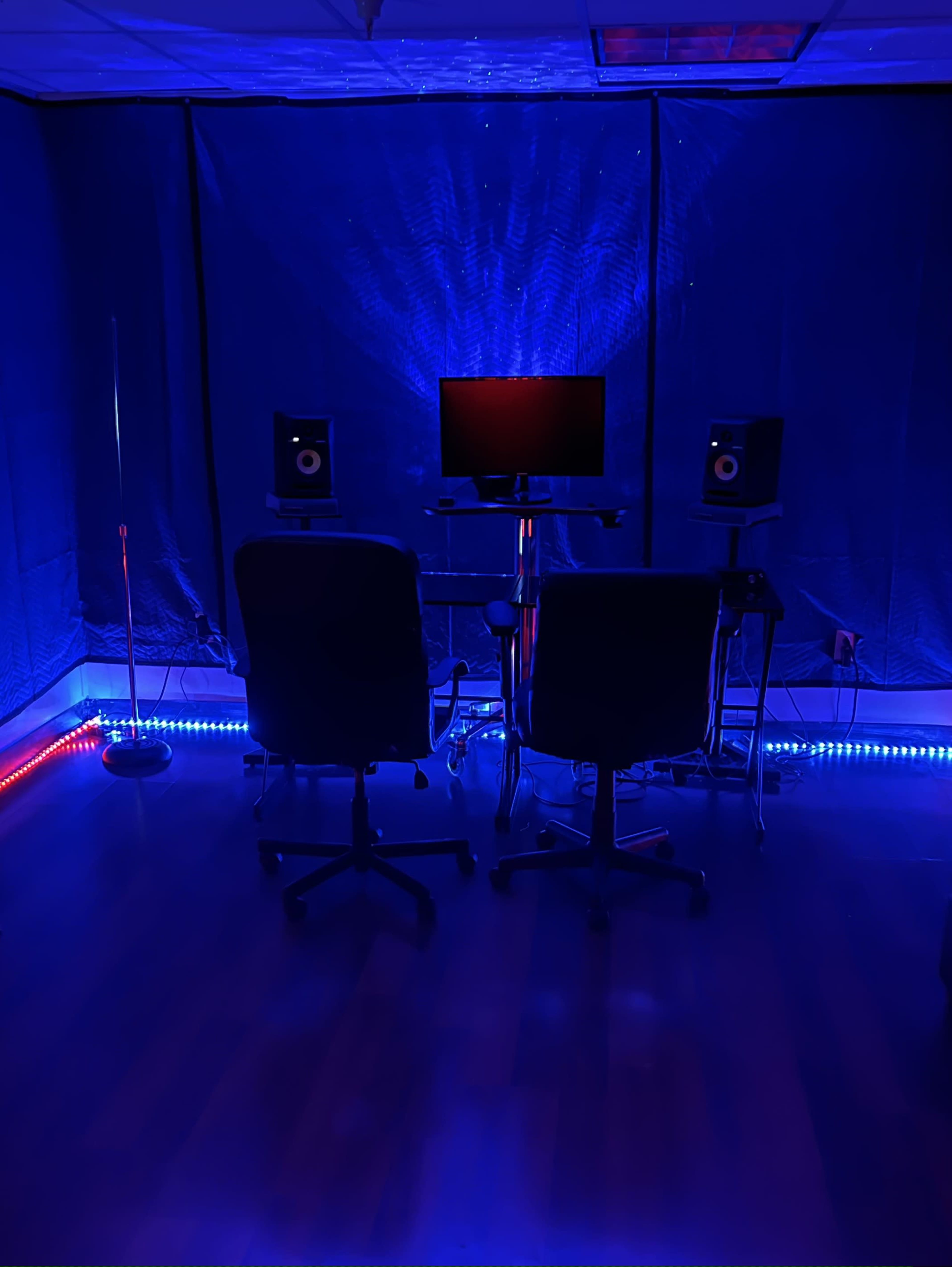 A dimly lit room with two black chairs, a computer monitor, and audio speakers, illuminated by blue lighting.