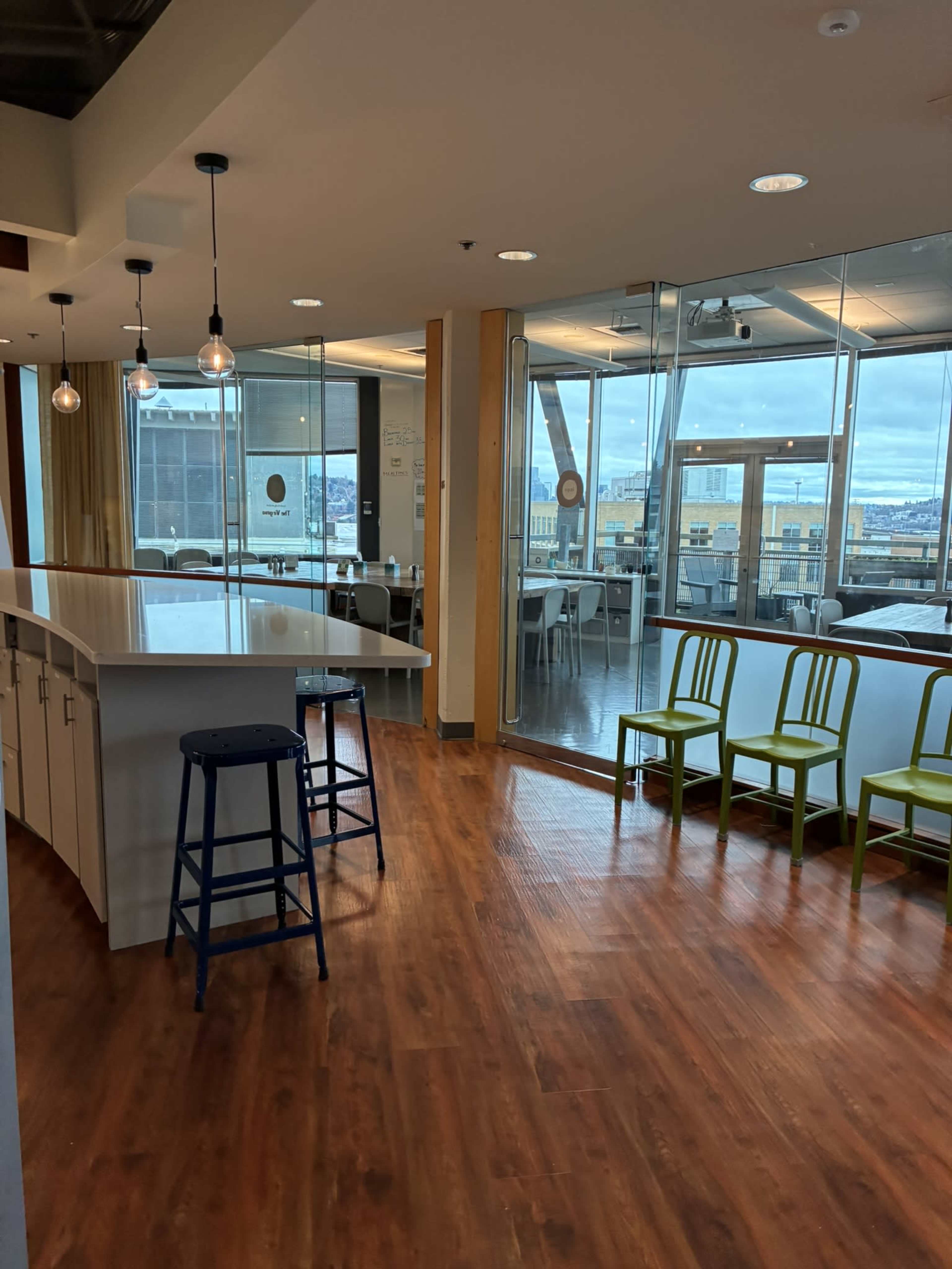 The image shows a bright, modern office space with a kitchen area featuring a large white countertop, barstools, and a row of green chairs along the wall, all with large windows offering a view of the city.