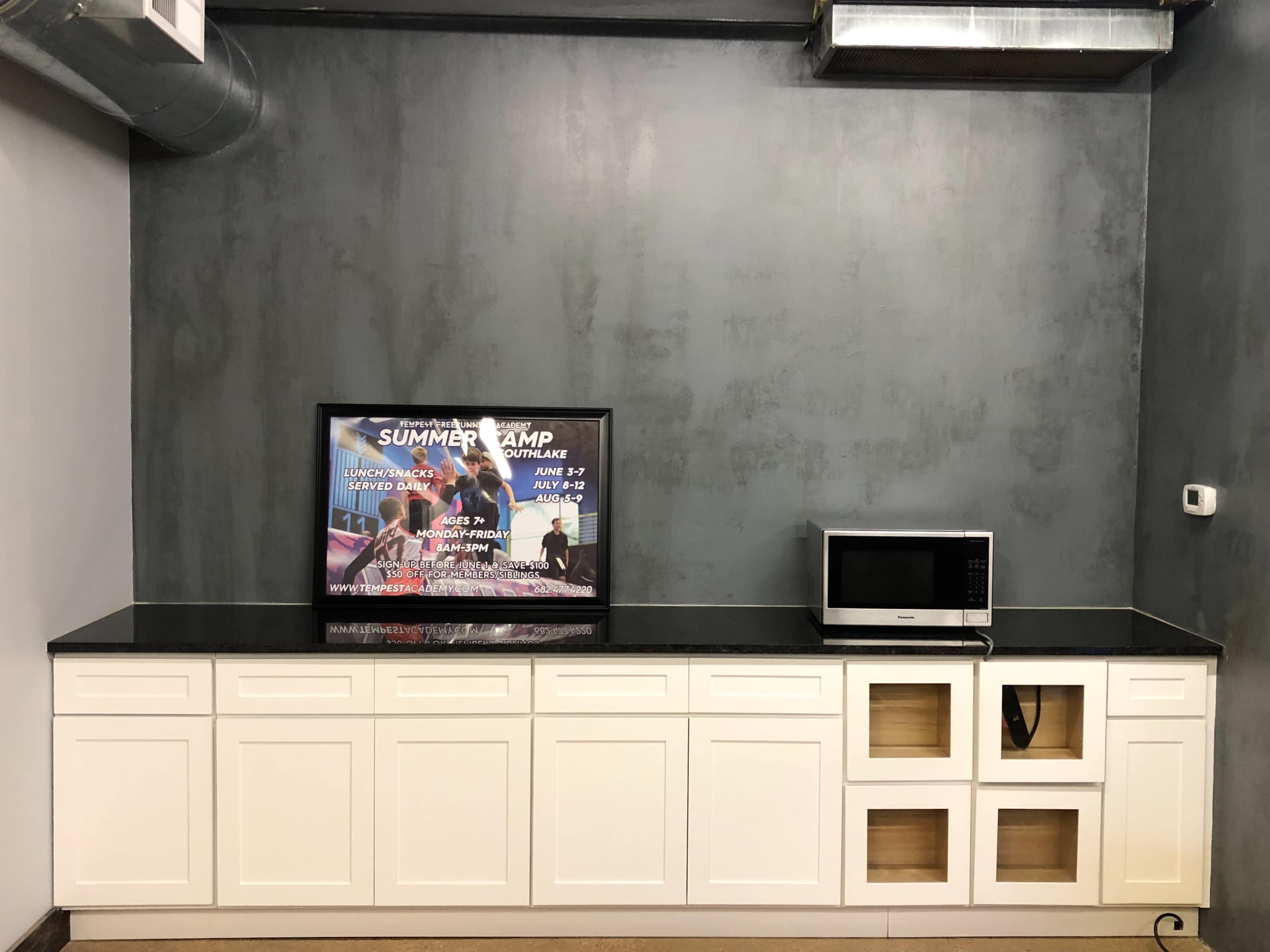 The image shows a kitchen area with a countertop, a microwave, and a television displaying a poster, all set against a gray wall.