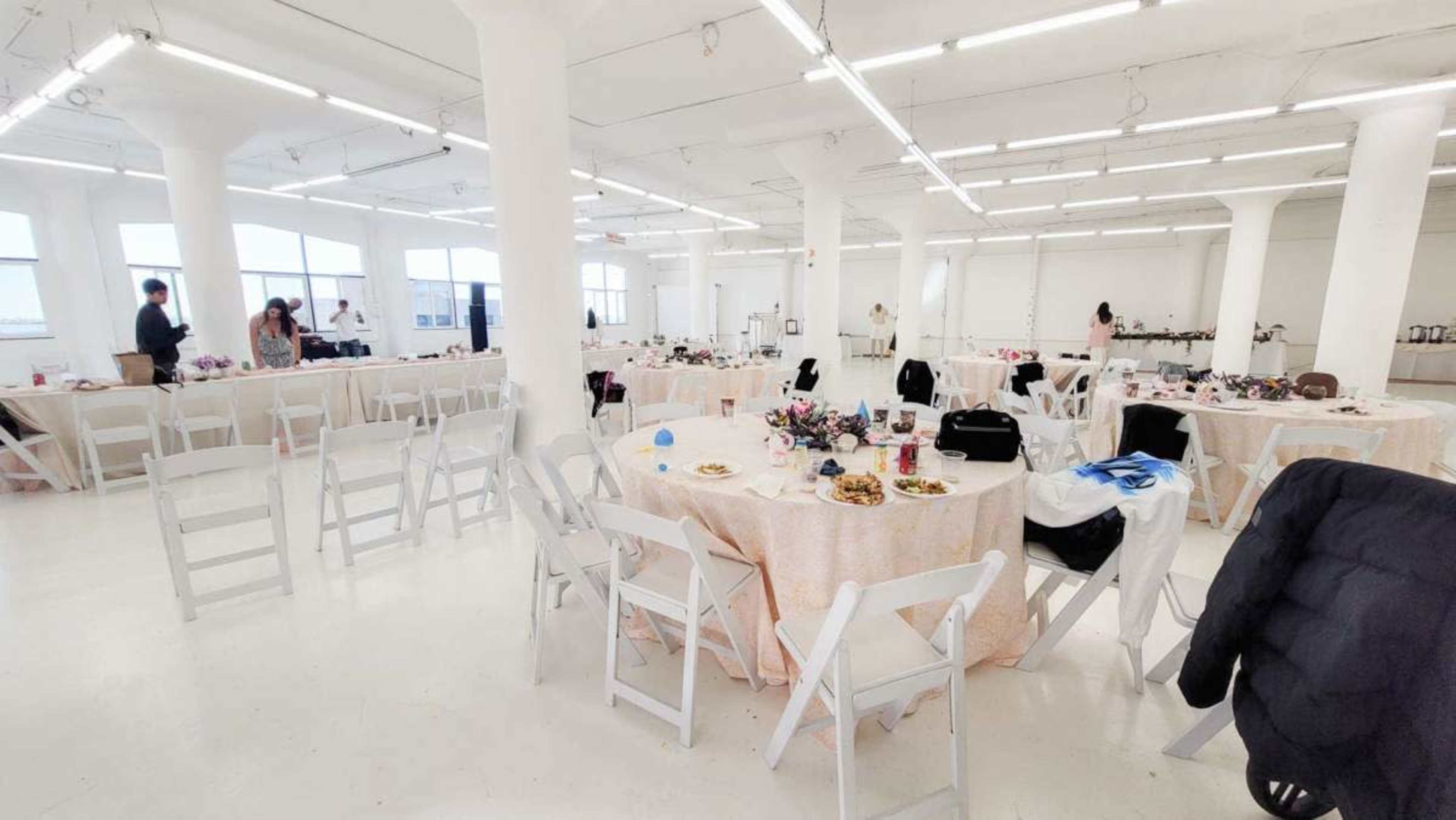 A spacious event room featuring several round tables set with light pink tablecloths, arranged with food and floral centerpieces.