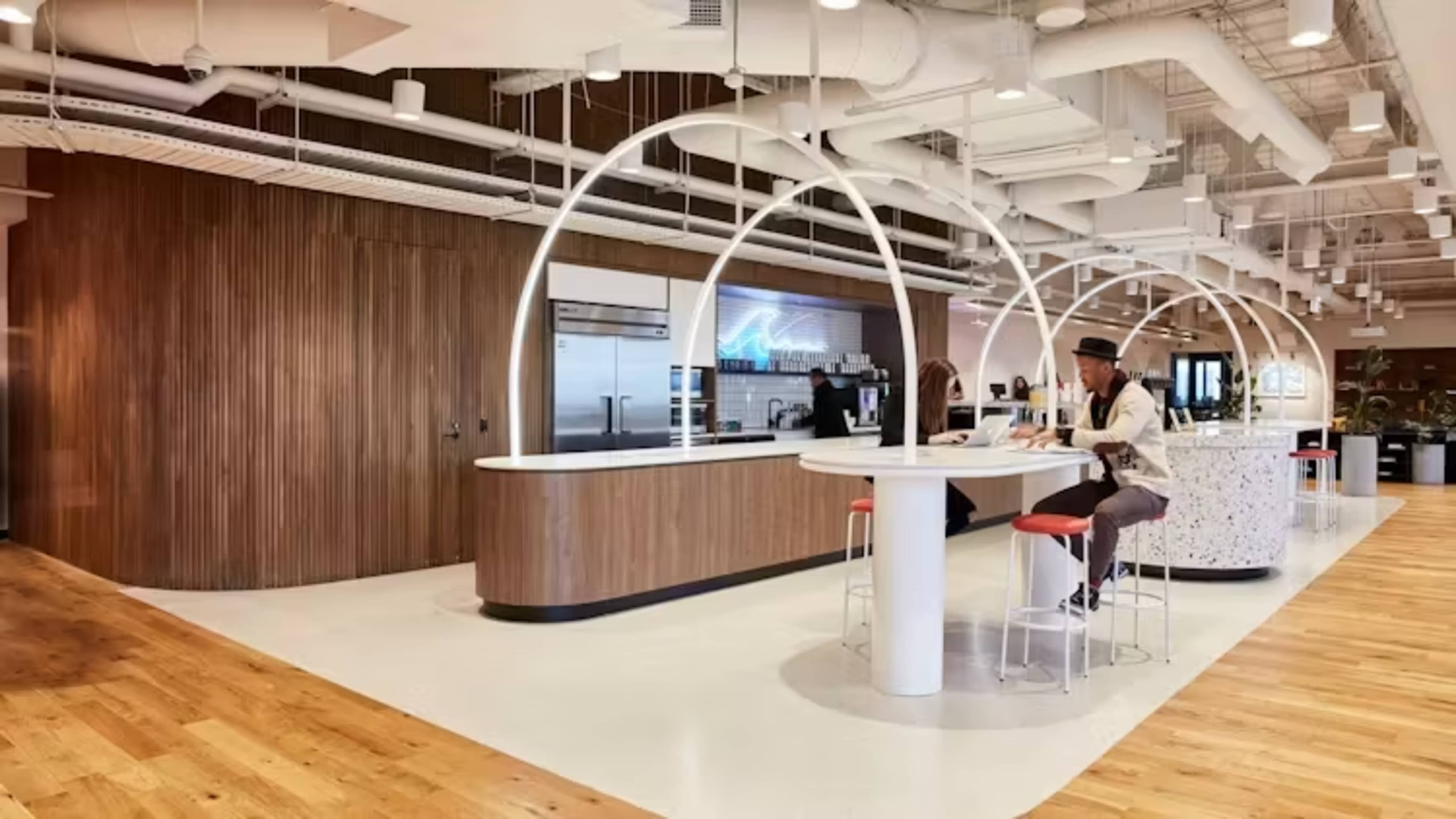 A modern office kitchen area features a sleek design with a large circular bar and tidy food preparation space, complemented by wooden accents and bright lighting.