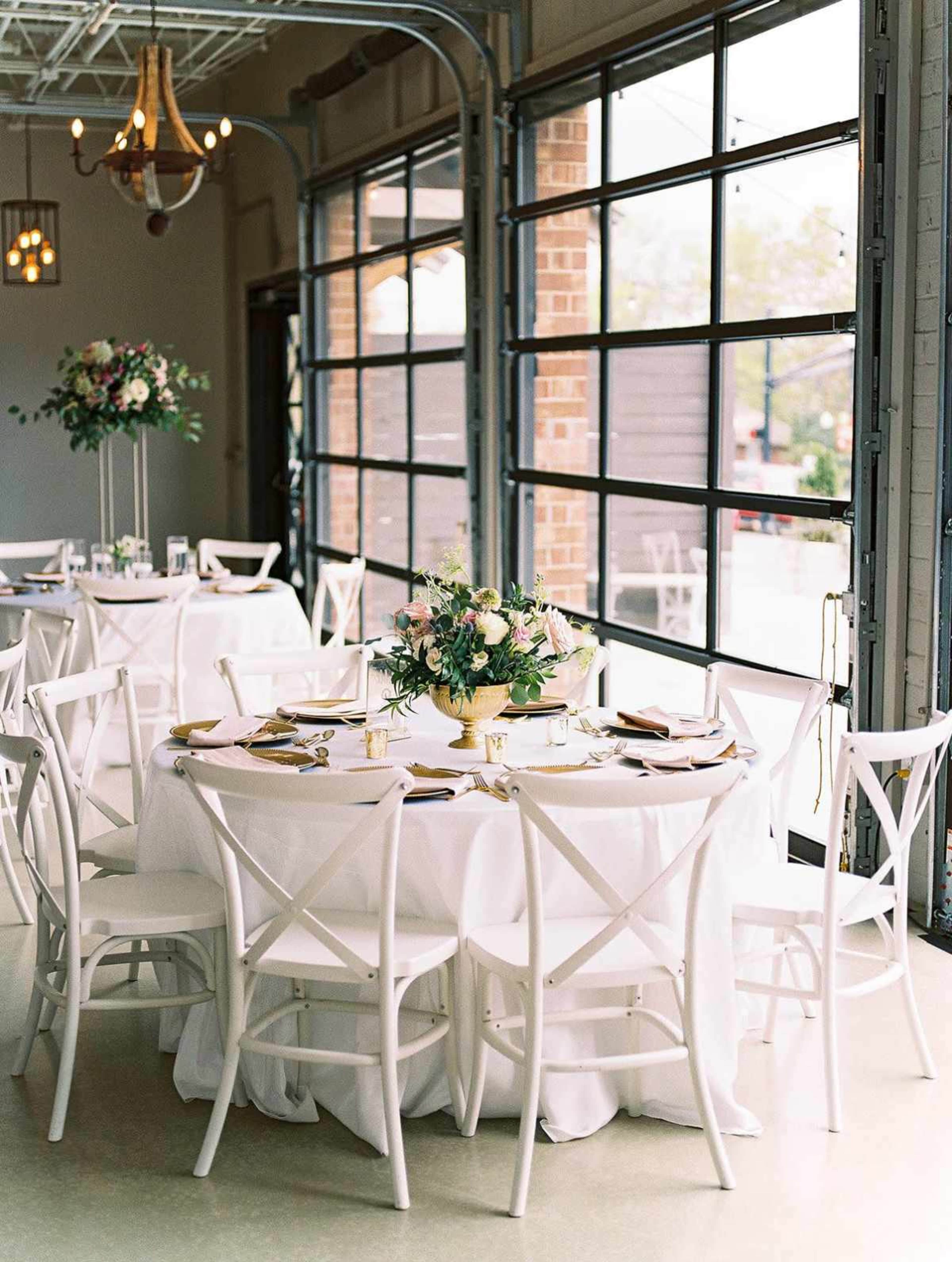The image shows a dining area with elegantly set round tables featuring white tablecloths and floral centerpieces, illuminated by natural light from large windows.