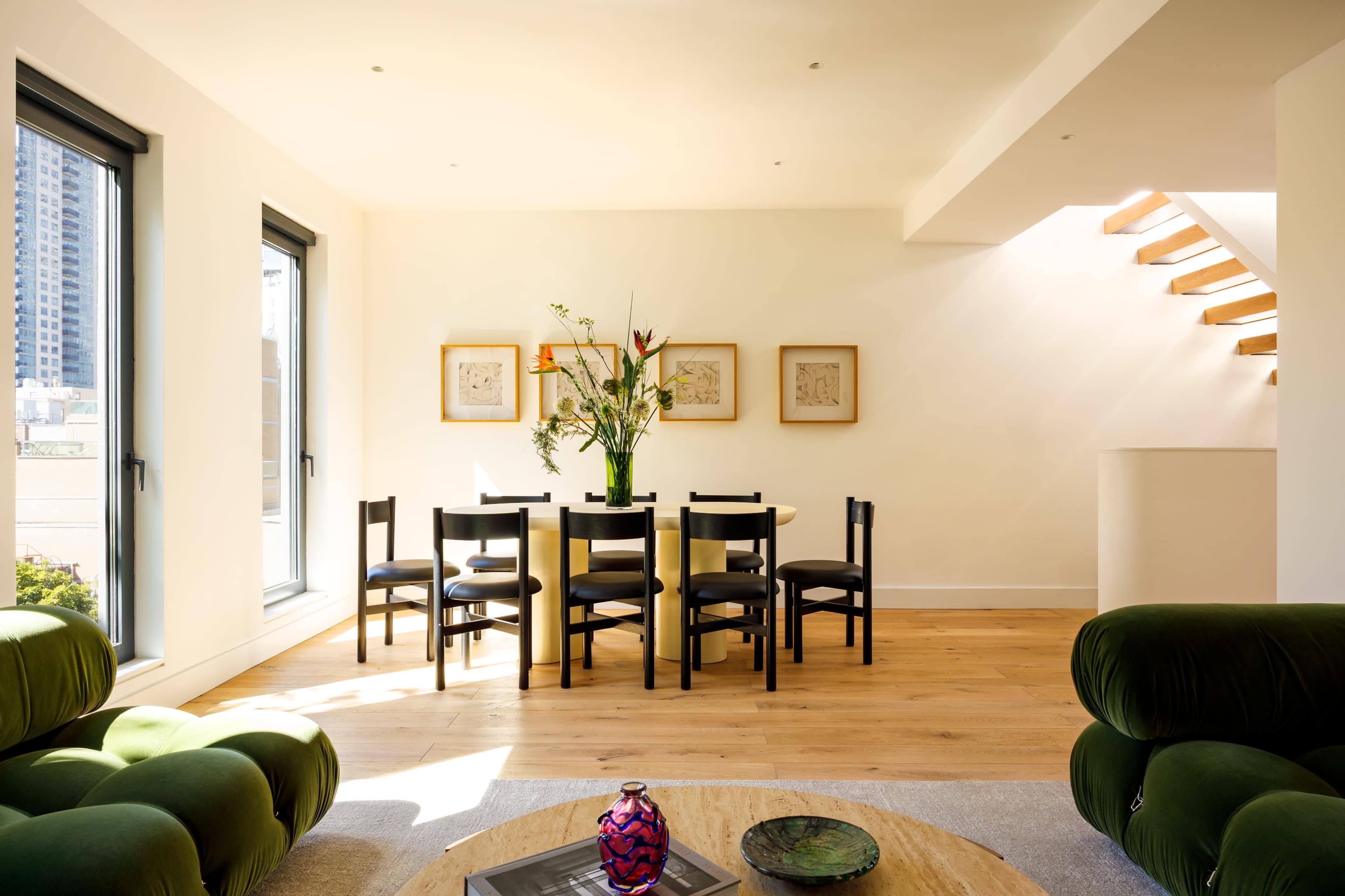 A modern dining area features a round table surrounded by black chairs, with a vase of flowers as the centerpiece and large windows letting in natural light.