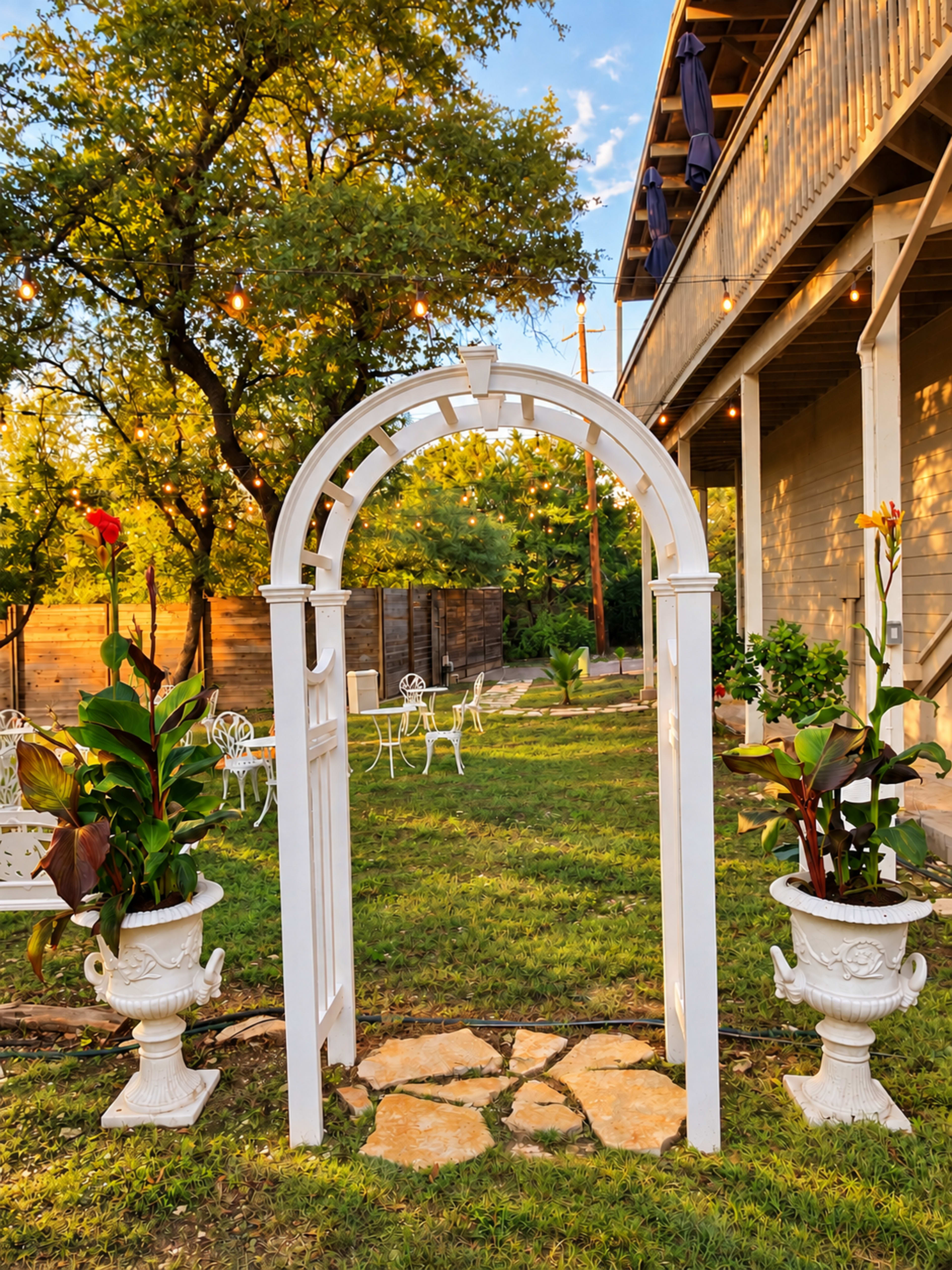 A white archway stands in a garden, flanked by potted plants, with a backdrop of trees and a building.