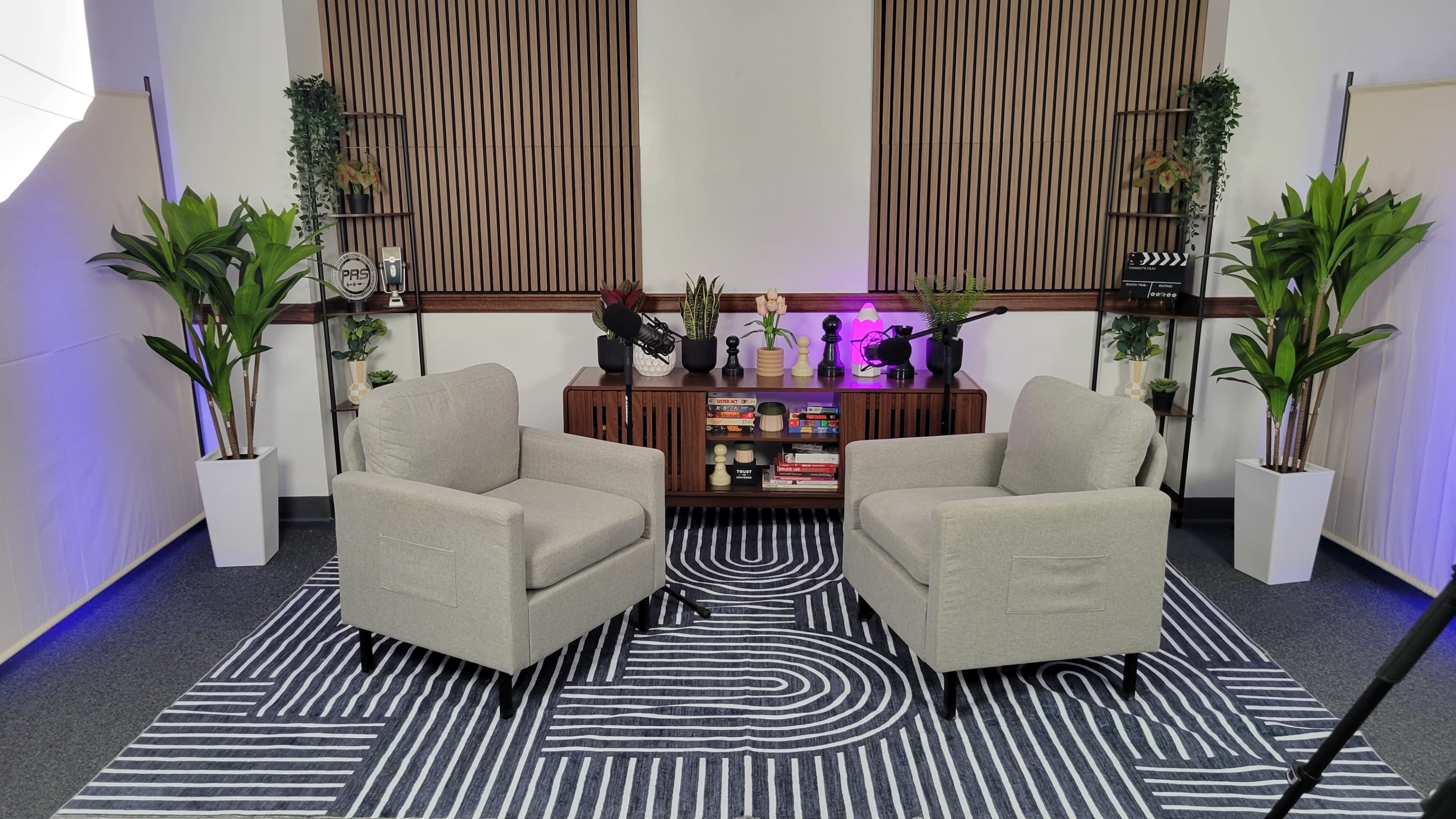 Two light-colored armchairs are positioned facing each other on a patterned rug in a well-decorated room featuring plants and a wooden backdrop.