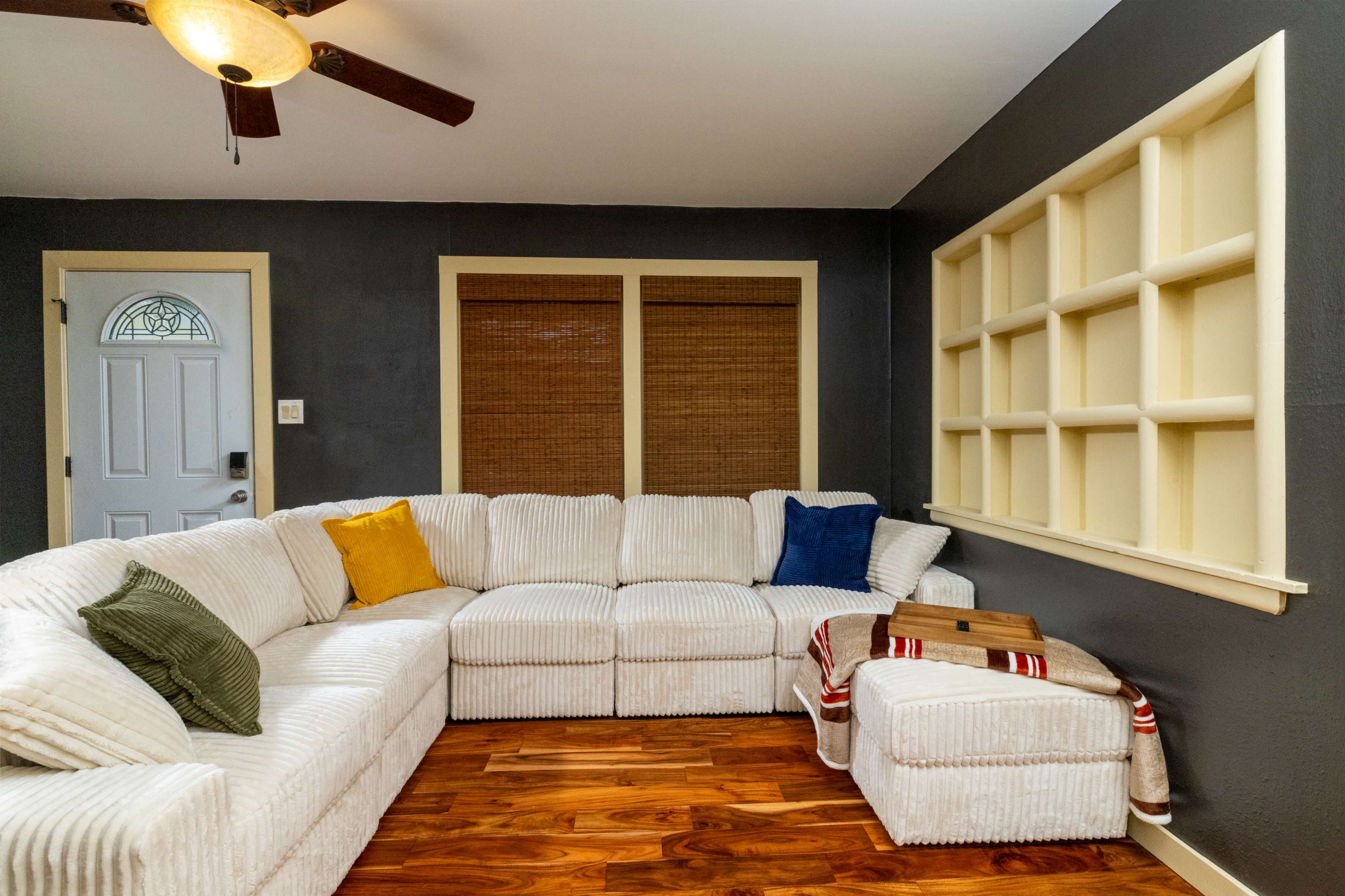 The image shows a living room featuring a large, white sectional sofa with accent pillows, a wooden floor, and a wall-mounted shelf by a door with blinds over the windows.