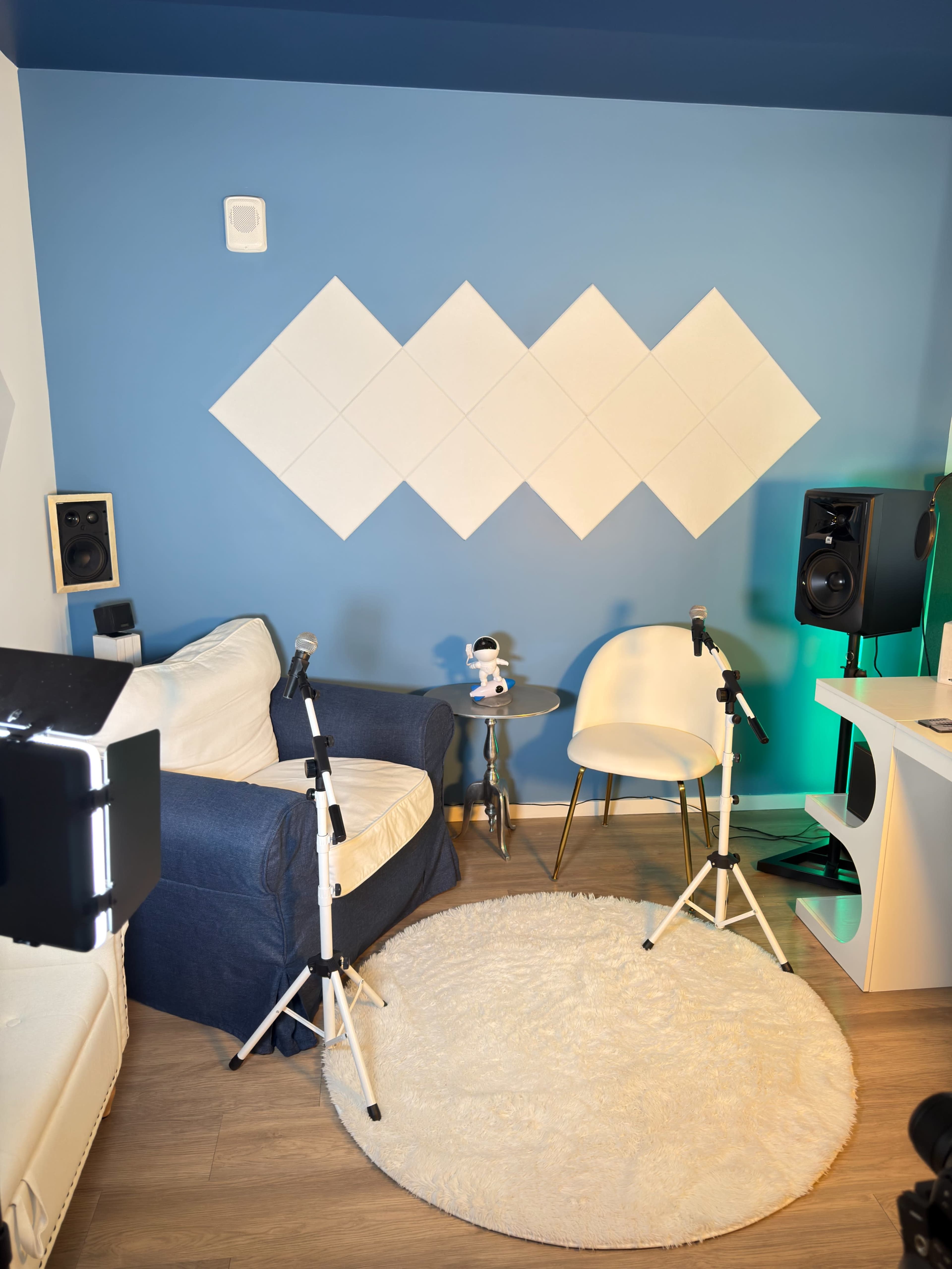 The image shows a small studio or recording space with two microphones on stands, a blue wall with decorative panels, and a cozy setup that includes a white chair and a plush rug.