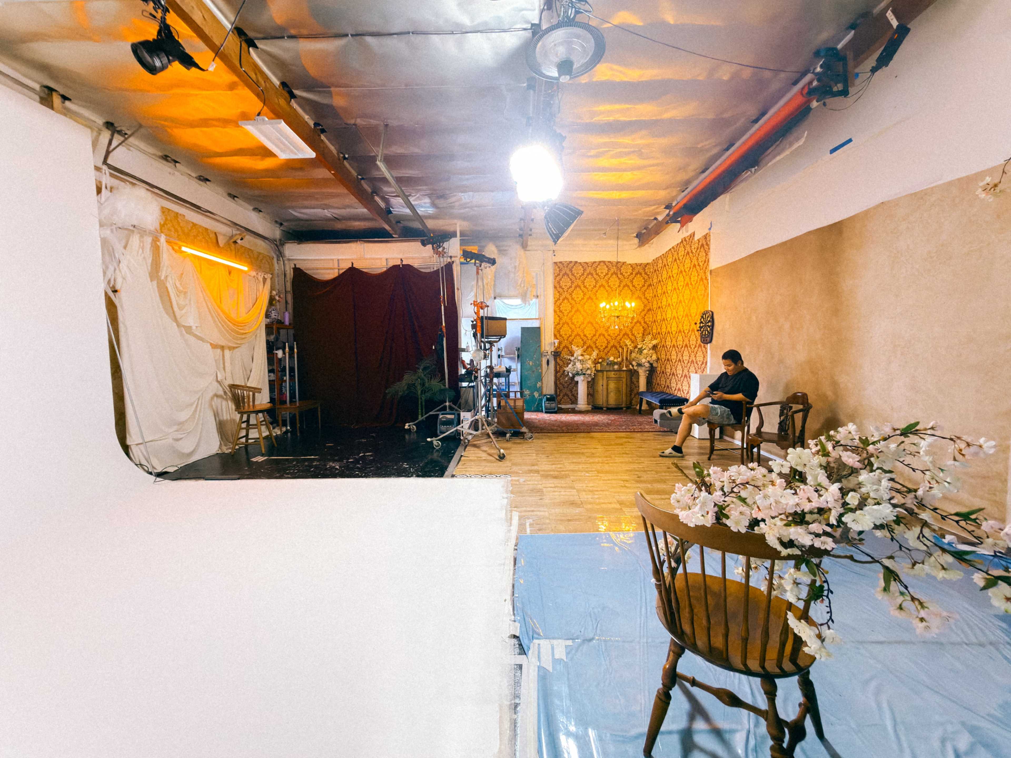 The image shows a photo studio with different backdrops, lighting equipment, a seated person, and a flower arrangement on a table.