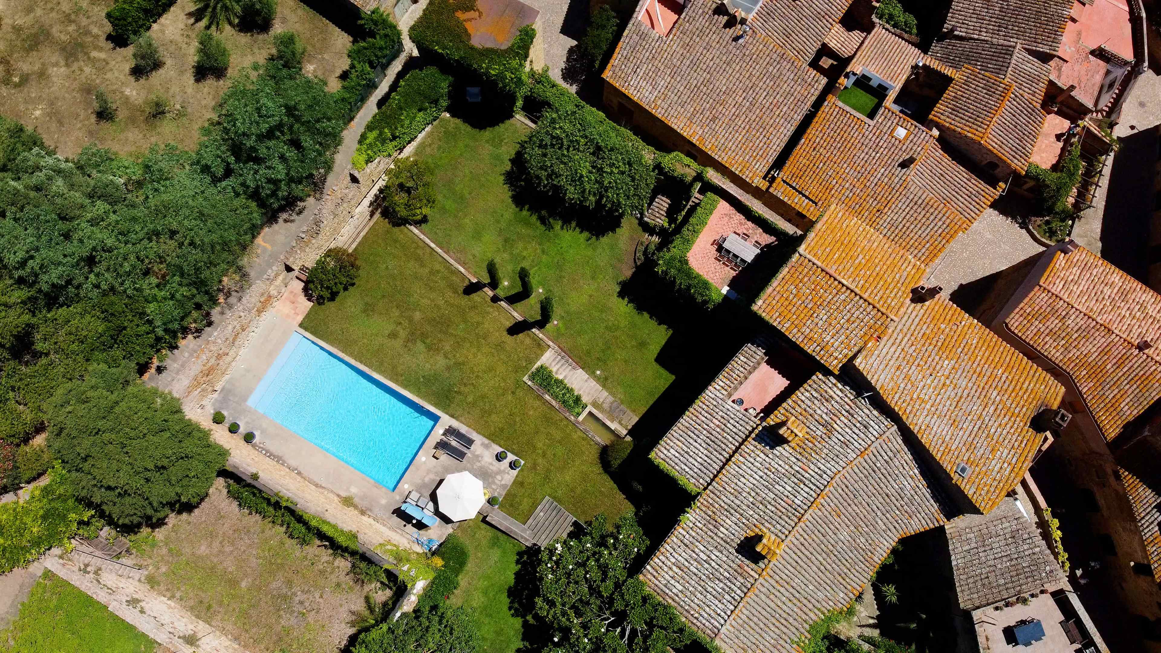 An aerial view shows a rectangular swimming pool surrounded by well-maintained grass and several houses with orange rooftops.