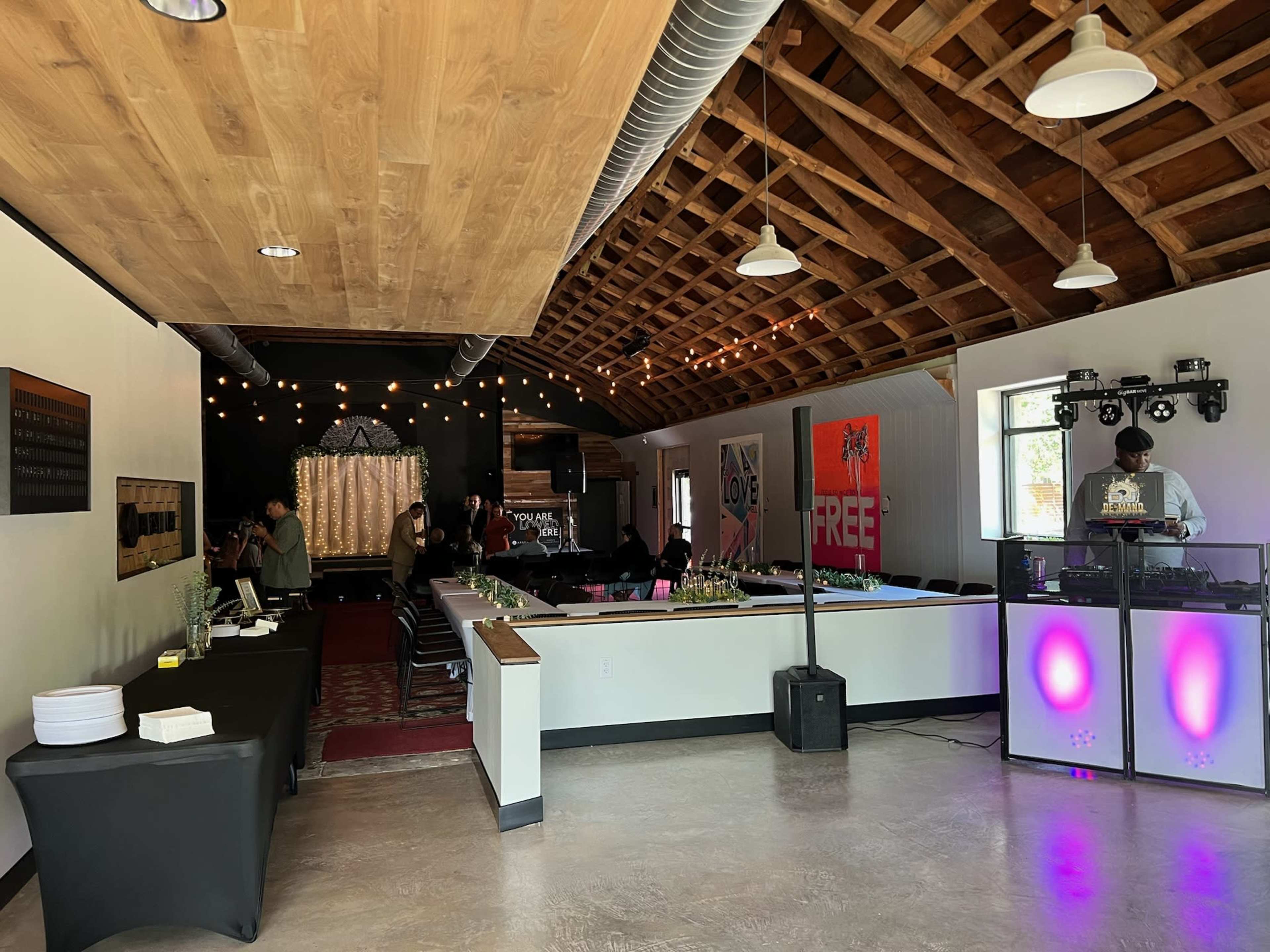 The image shows an indoor venue with a wooden ceiling, a DJ setup in one corner, and tables arranged for an event.
