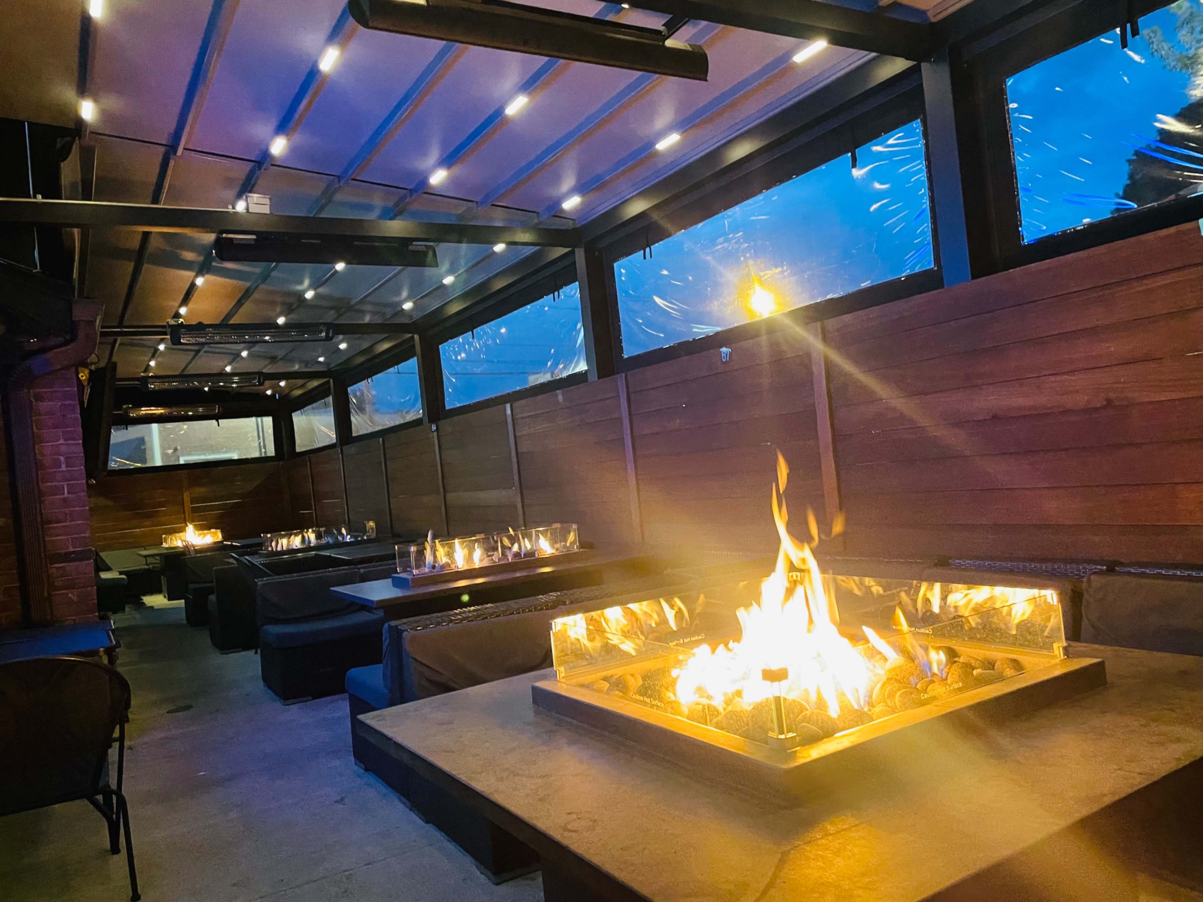 The image shows a modern, covered outdoor seating area with several fire pits and a view of a rainy evening outside.