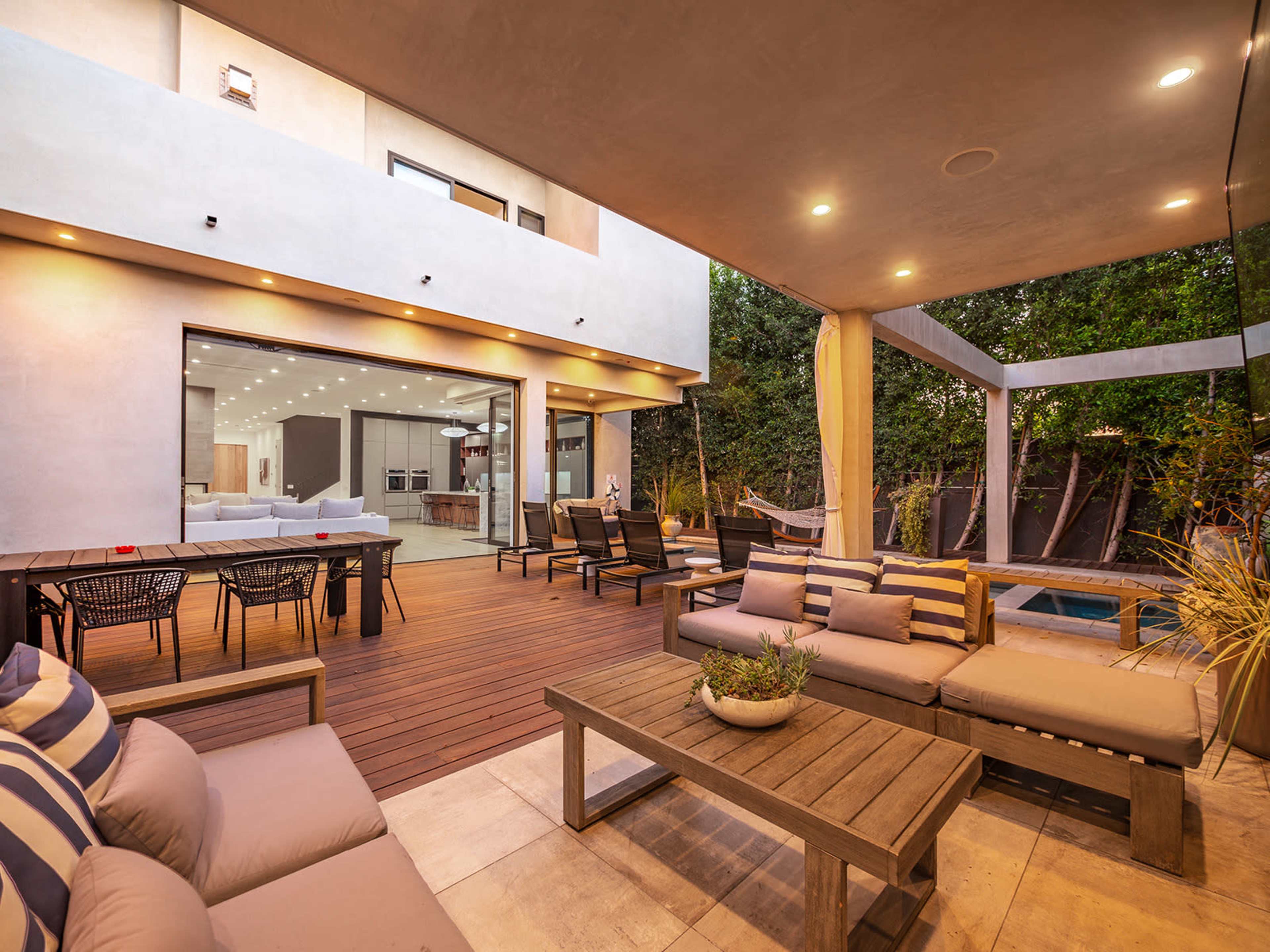 The image shows a modern outdoor patio area with wooden furniture, a lounging space, and an adjacent pool, surrounded by greenery.