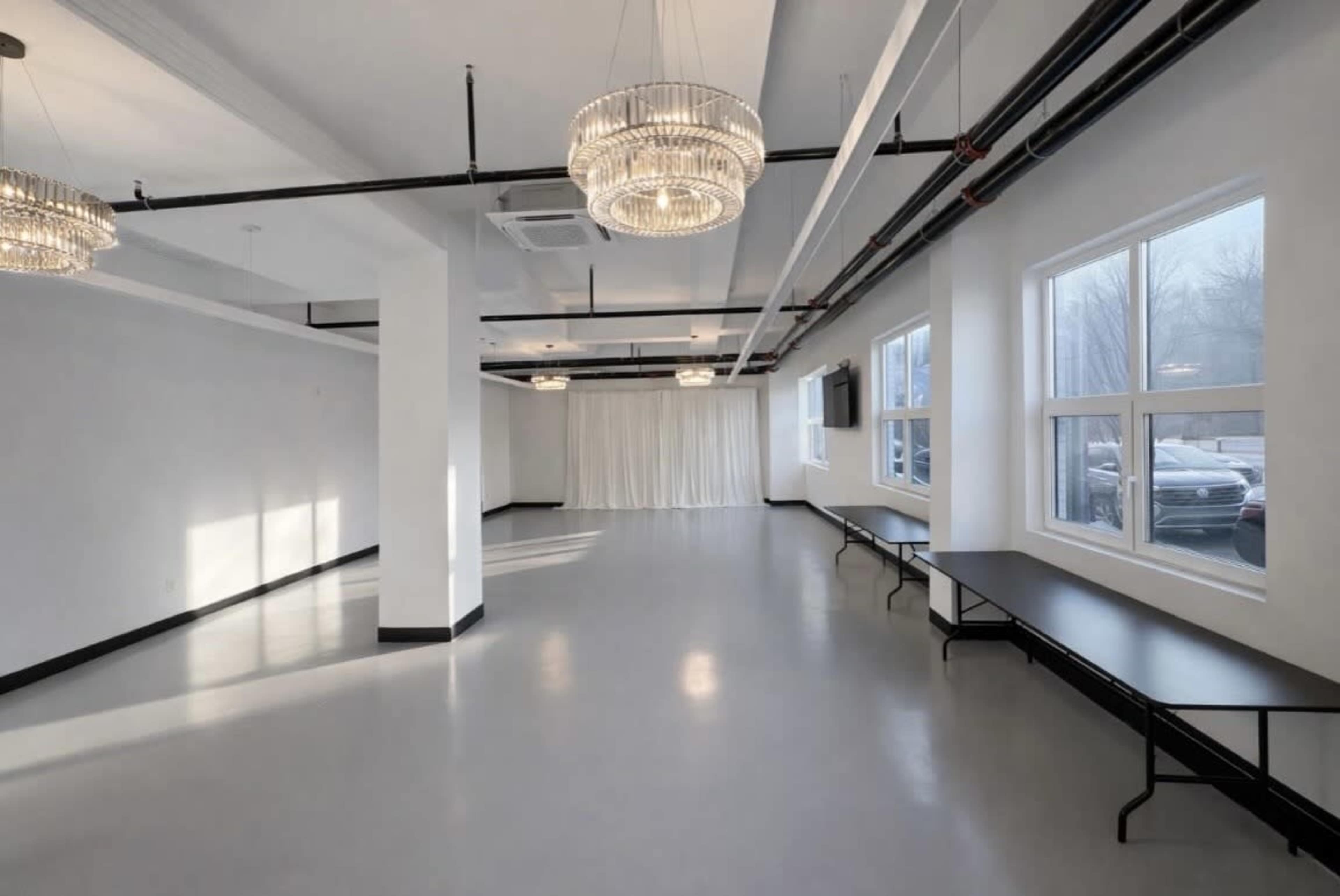 The image shows an empty, spacious room with gray floors, white walls, two chandeliers, and long tables along one side.