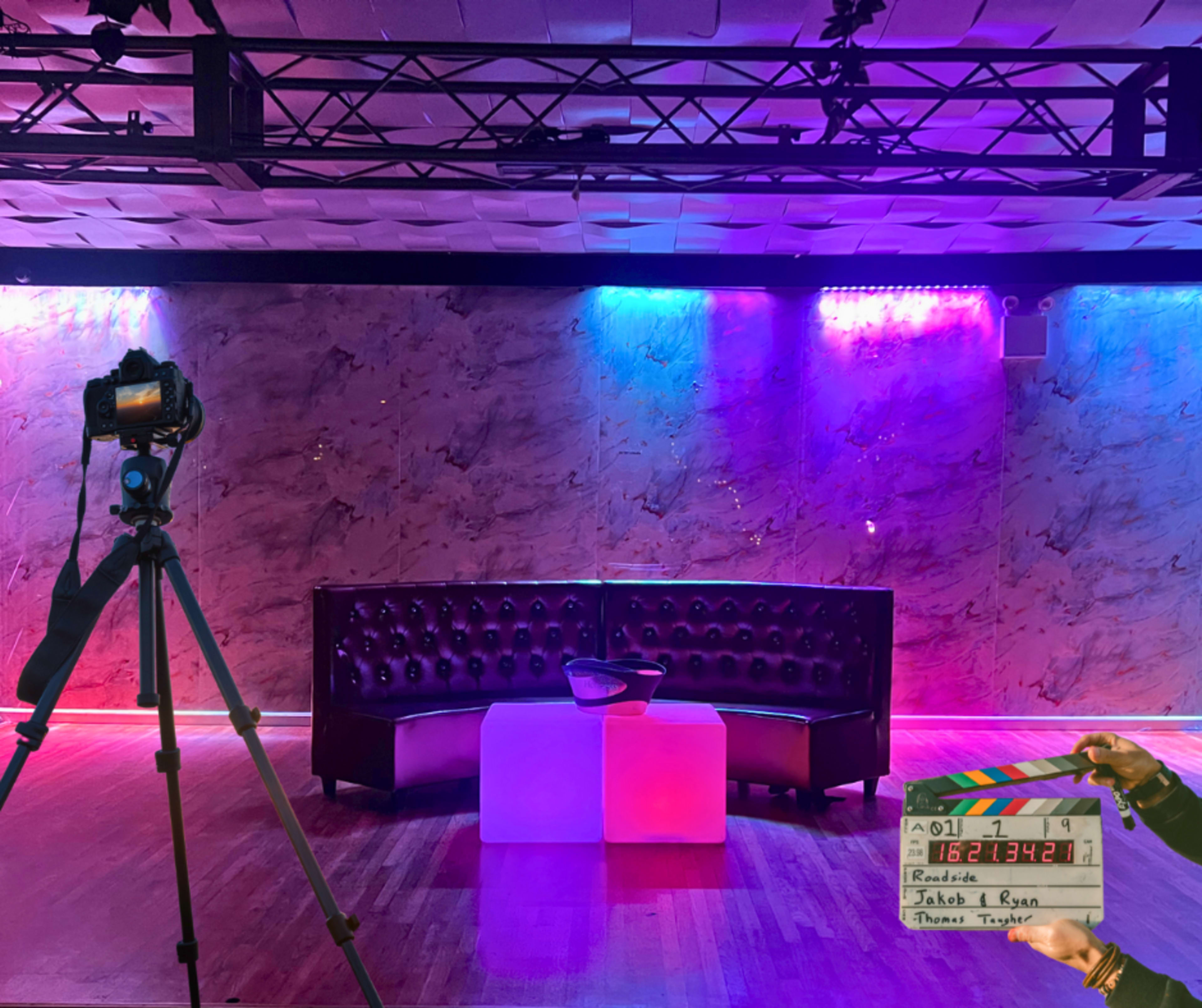 A camera tripod is set up in front of a modern, well-lit stage featuring a dark leather couch and two illuminated tables.