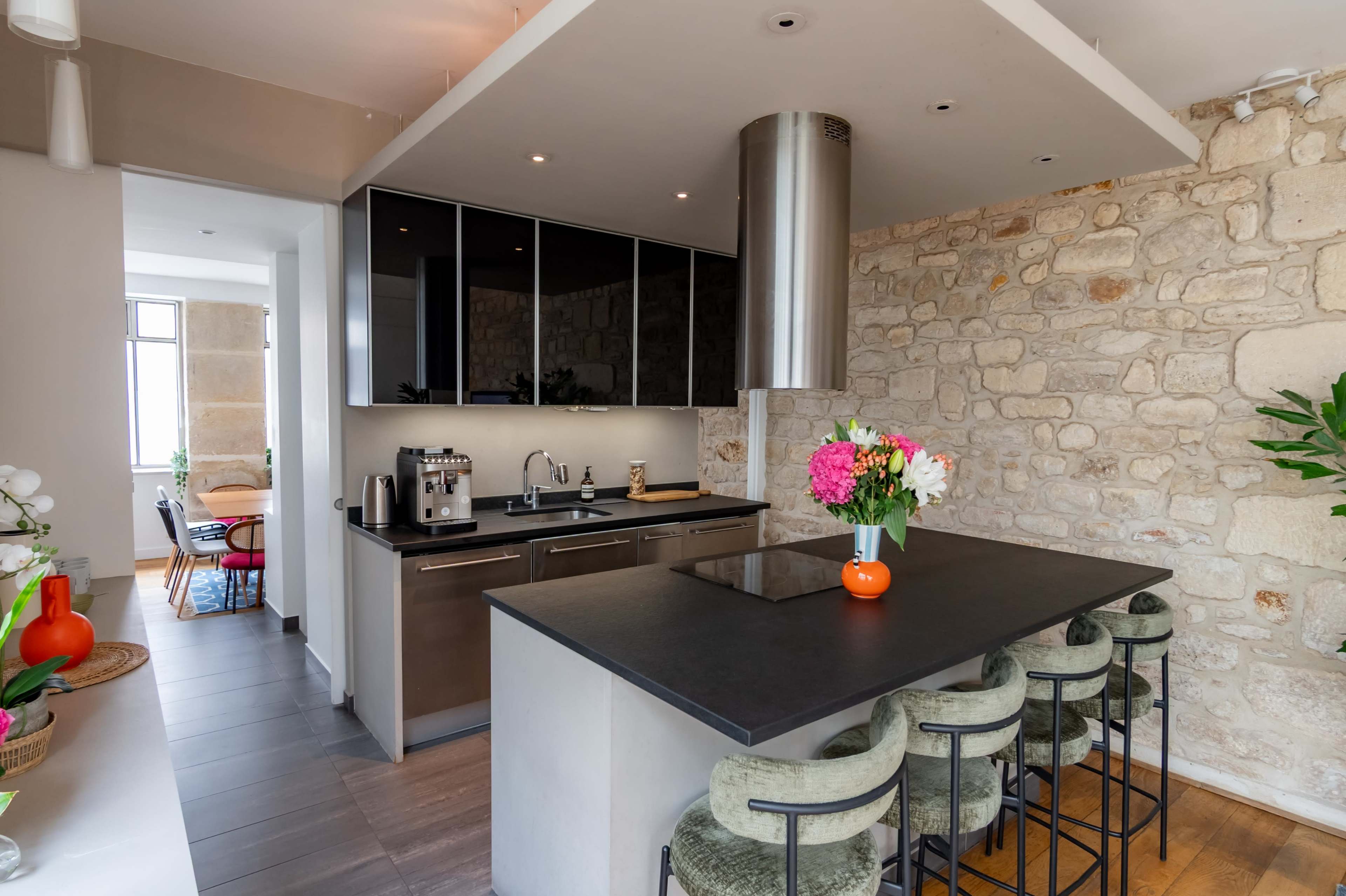 The image shows a modern kitchen with a sleek black countertop, a stainless steel range hood, and a stone wall, complemented by a dining area in the background.