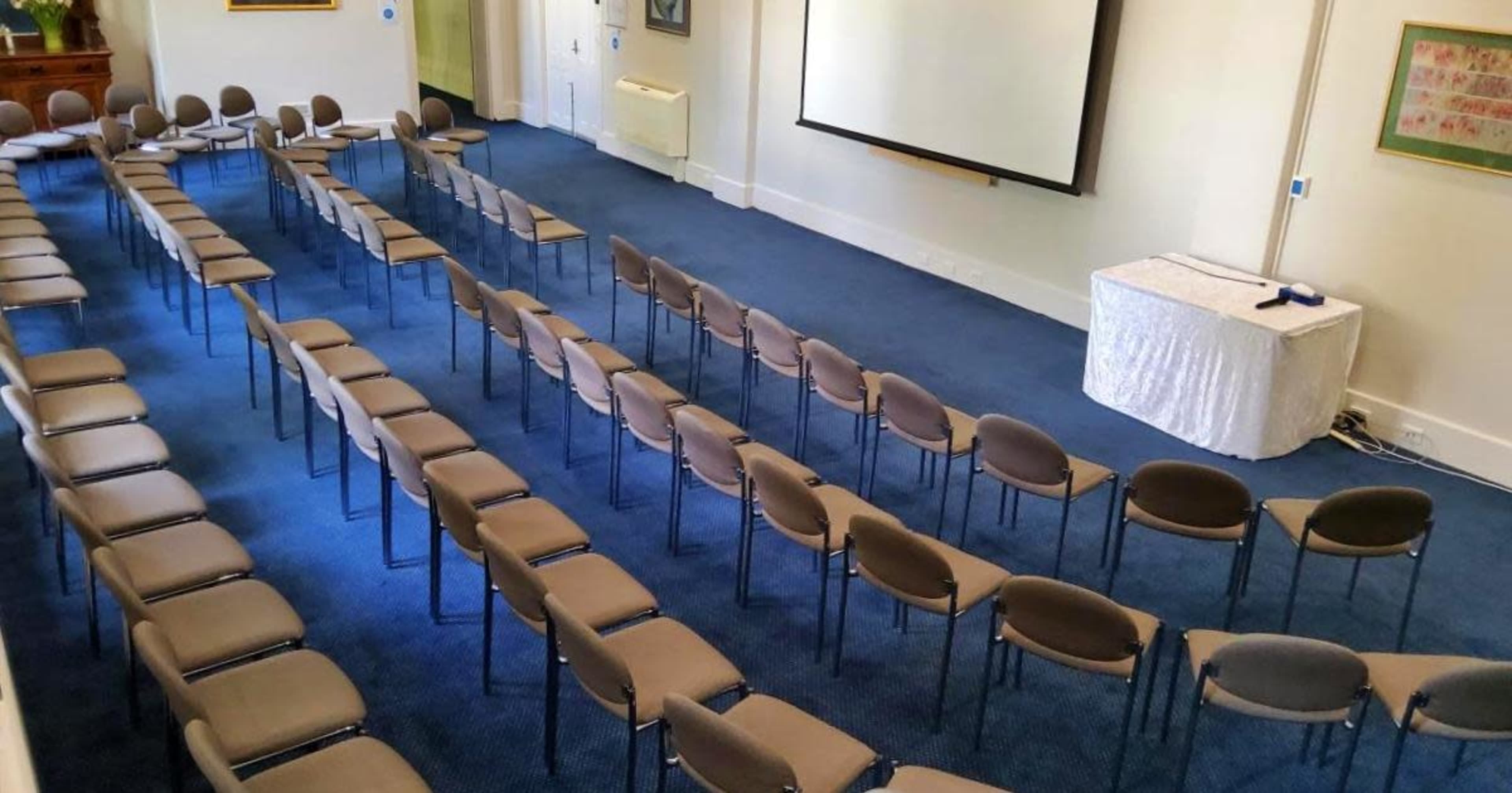 A room set up for a meeting with rows of chairs facing a blank screen and a small table at the front.