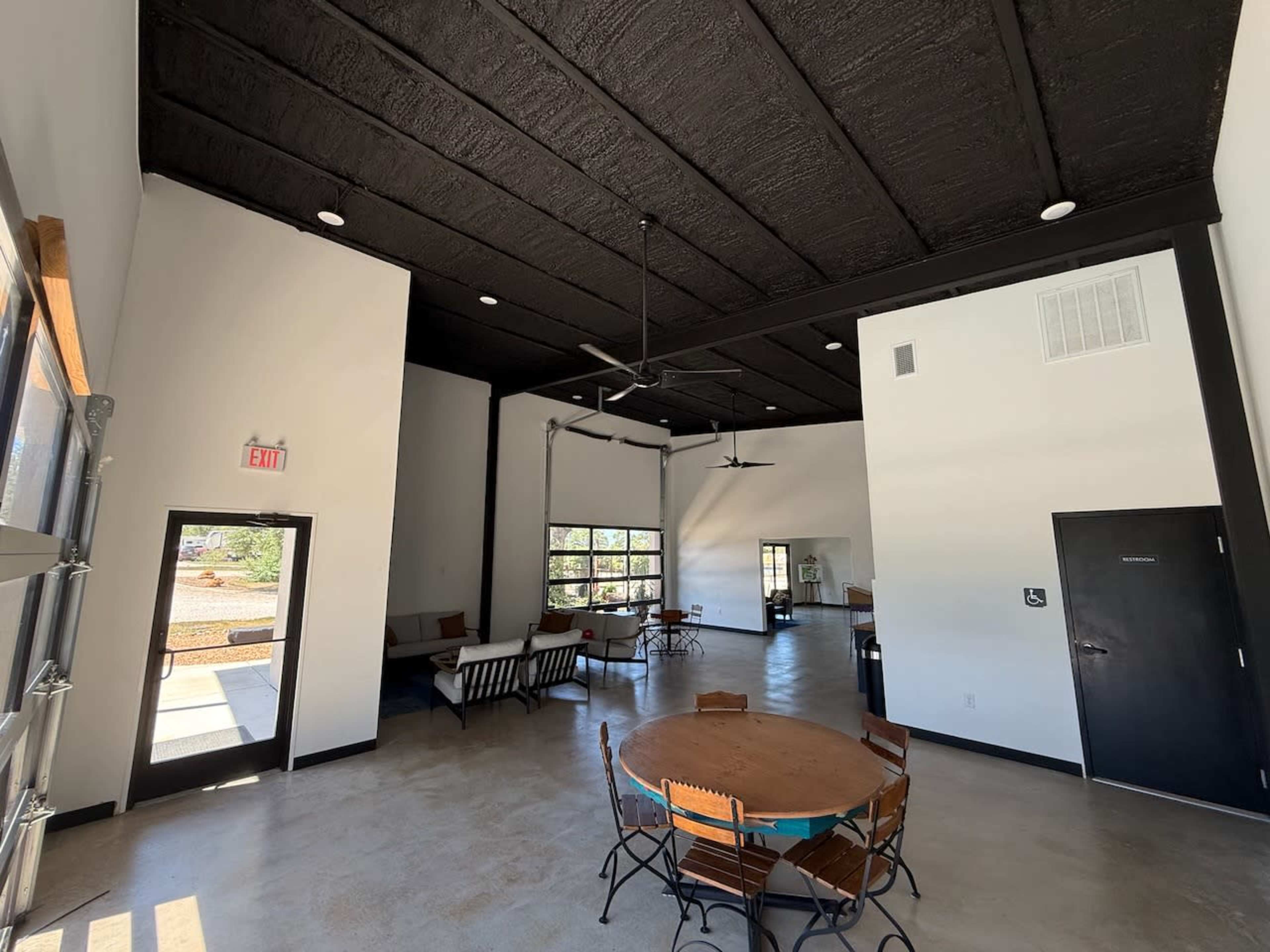 The image shows a spacious interior room with high black ceilings, large windows, and a mix of seating areas, including a round wooden table with chairs, sofas, and a door leading outside.