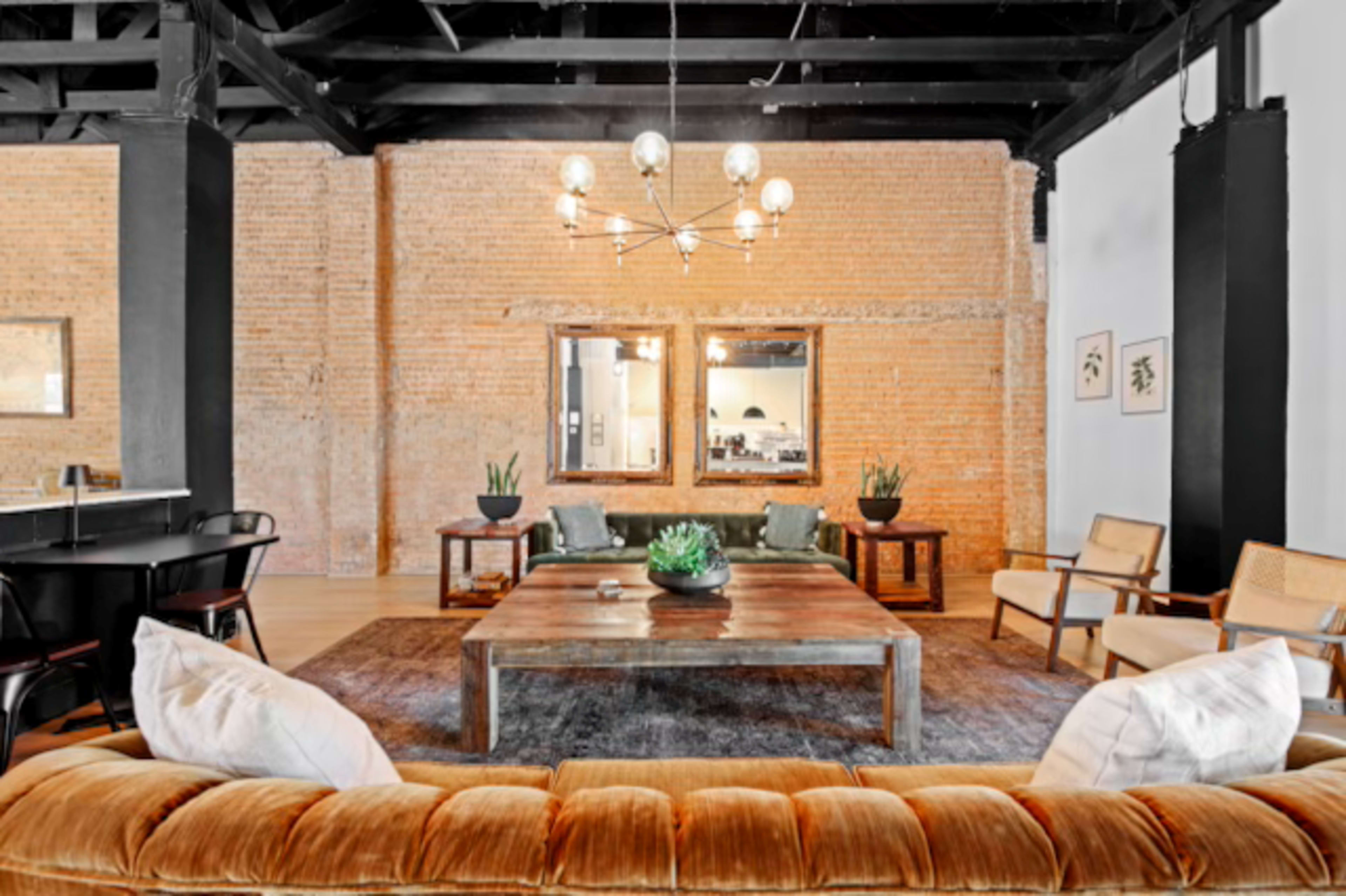 The image shows a spacious living area featuring a large wooden coffee table, a velvet sofa, and several plants, with exposed brick walls and industrial lighting fixtures.