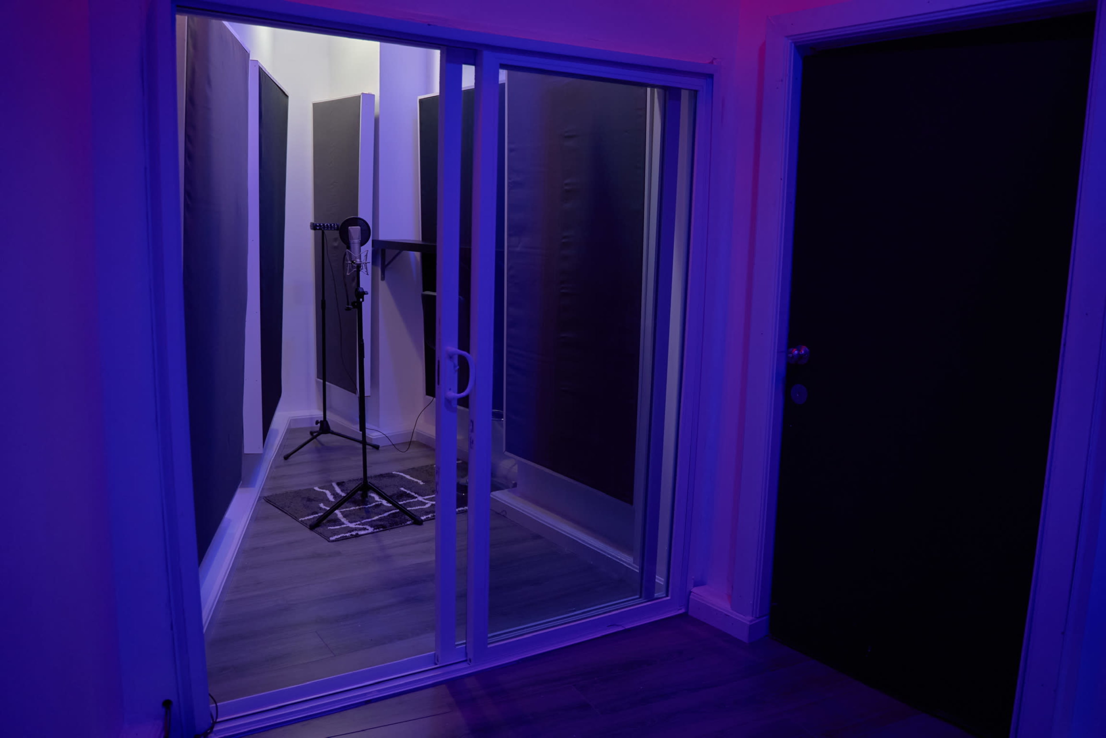 The image shows a soundproof recording studio viewed through a glass door, with acoustic panels lining the walls and a microphone setup inside.