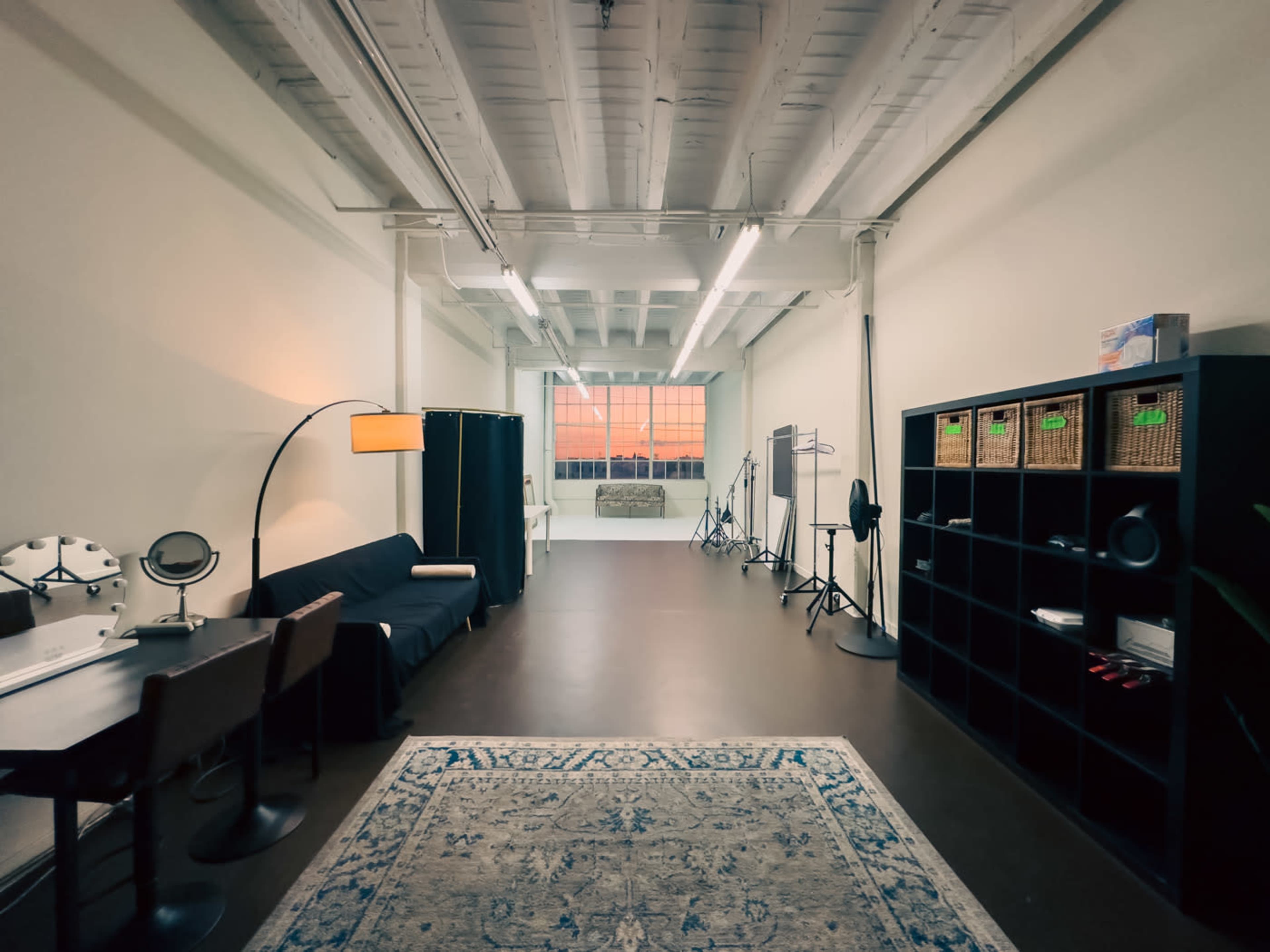 The image shows a spacious, well-lit room with a sofa, chairs, a mirror, and a shelving unit, featuring large windows that reveal a sunset outside.