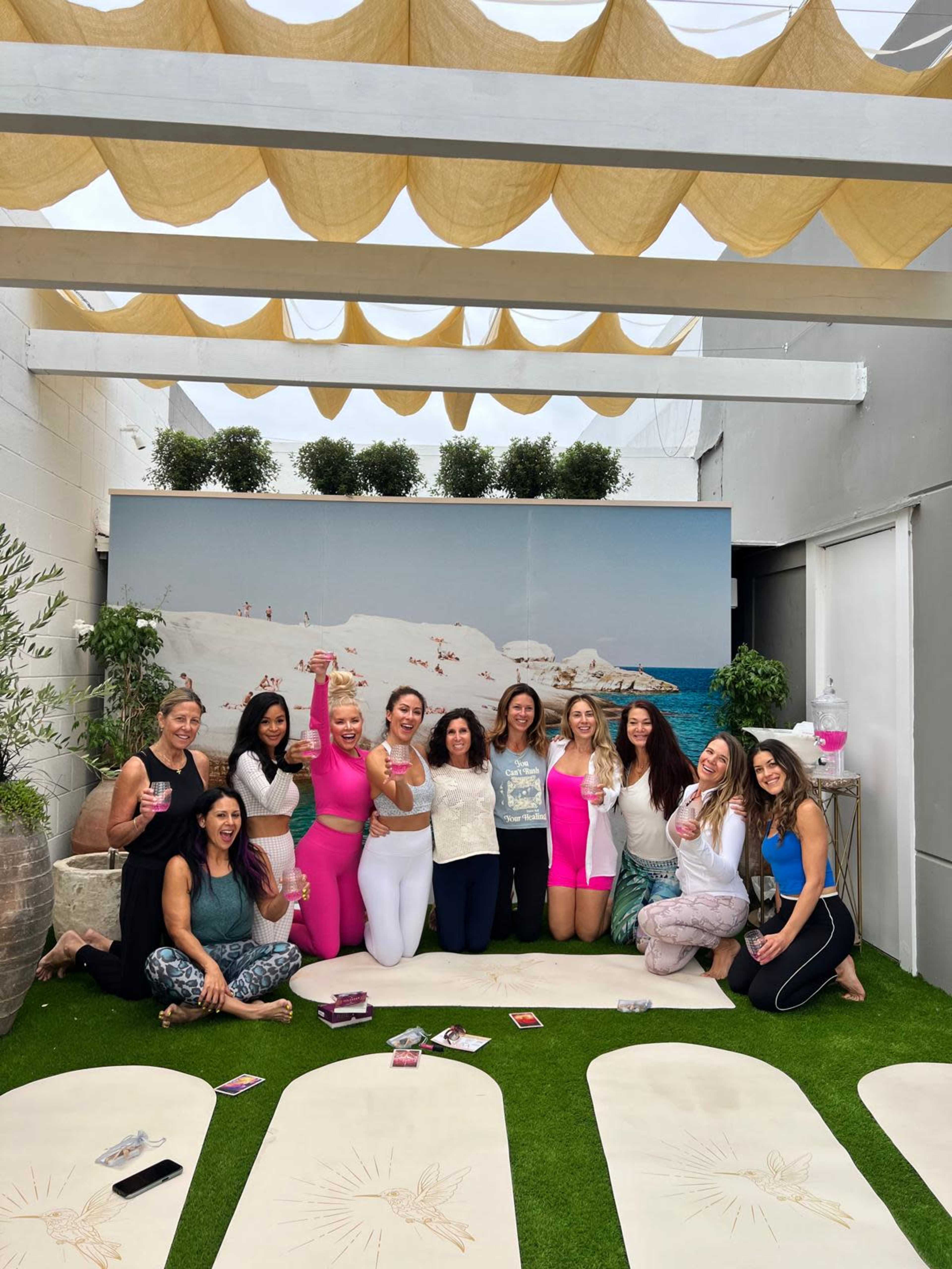 A group of women poses together in a yoga studio decorated with plants and a beach backdrop, with yoga mats spread out on the green turf.