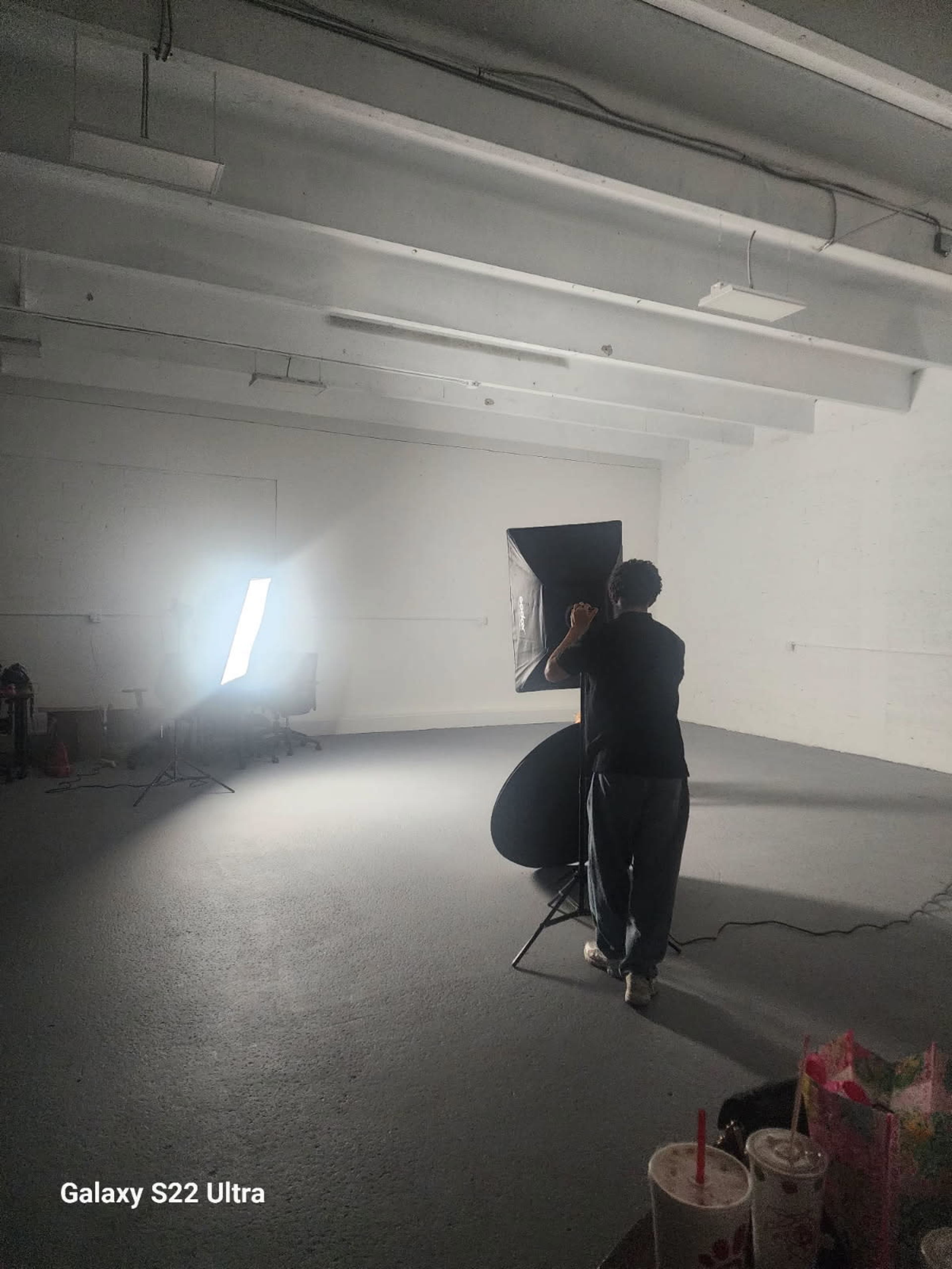 A person adjusts a light setup in an empty room with a white wall and concrete floor.